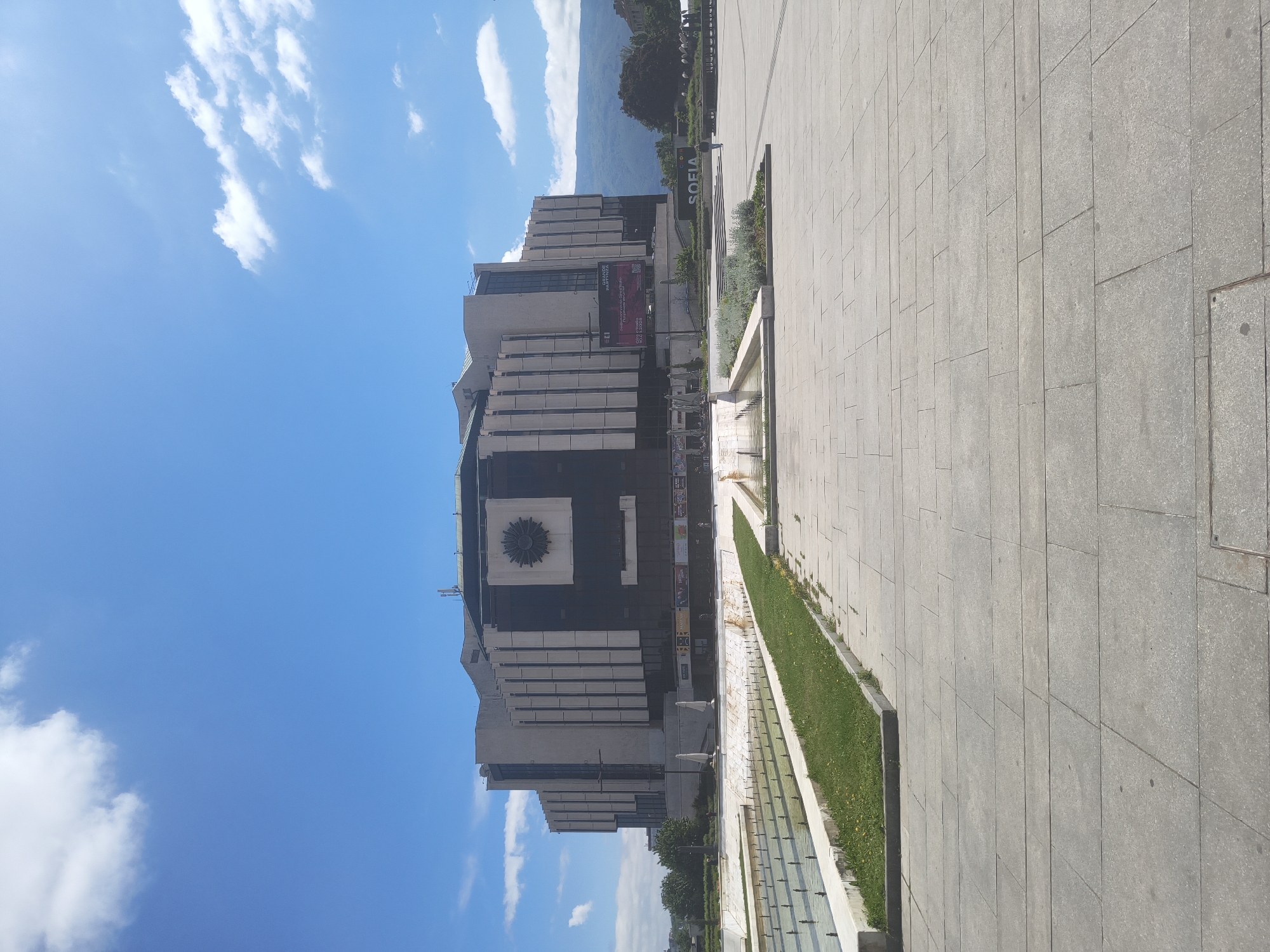 The image shows the National Palace of Culture in Sofia, Bulgaria. The building has a modernist architectural style with a prominent dark square with a sunburst design on its facade. There is a large open square paved with grey tiles in the foreground, extending towards the right. A long, narrow water feature runs parallel to the building, lined with strips of grass. In the background, behind the building, are trees and a view of mountains in the distance. The sky is bright blue with scattered white clouds. A sign with the word "SOFIA" is also visible. The photograph appears to have been taken during the day in good weather.