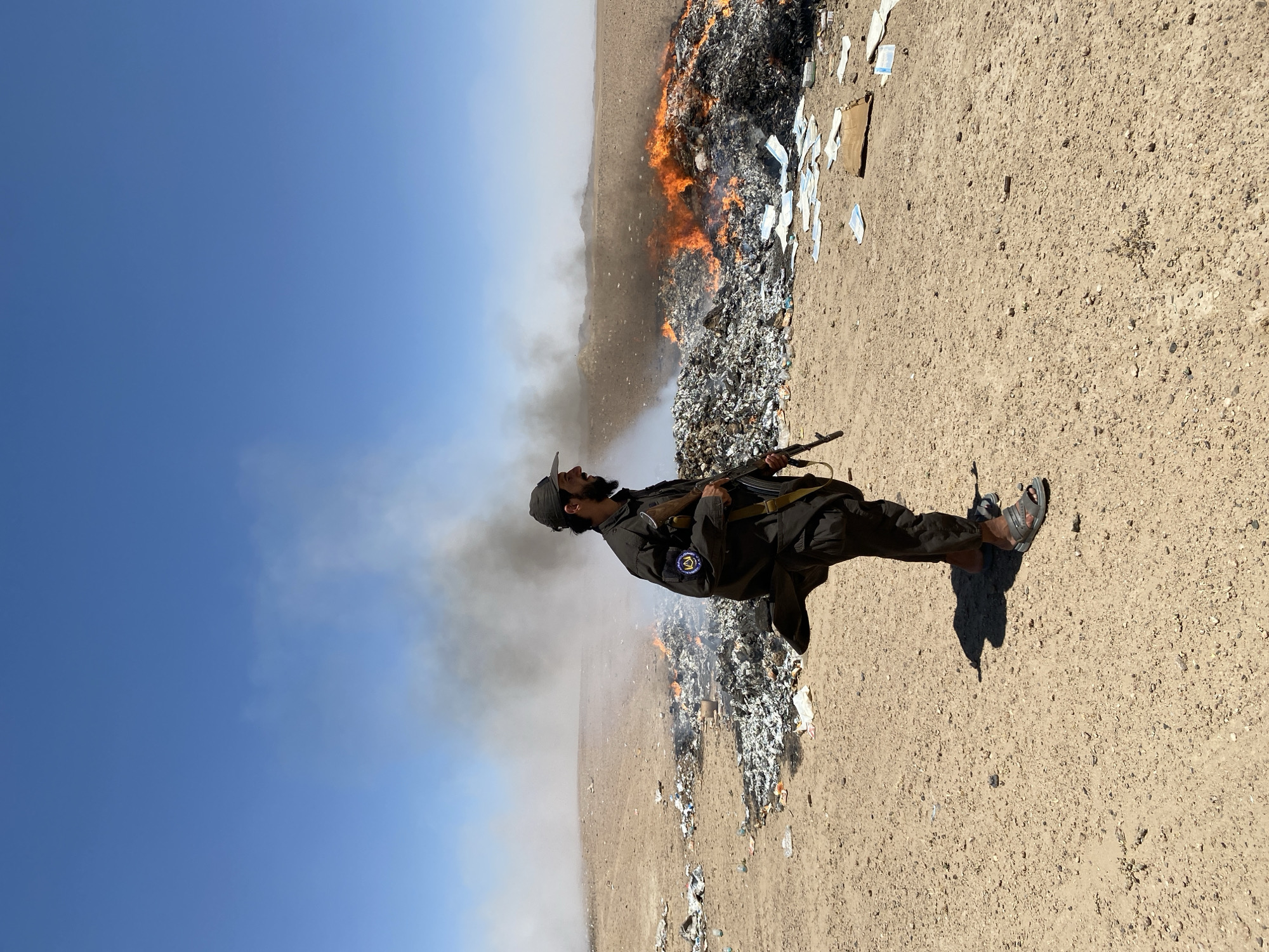 A man wearing a dark uniform and a baseball cap stands in a dry, barren landscape with a clear blue sky overhead. He is holding a rifle. To his left, a large pile of trash is burning, with flames and smoke billowing upwards. The ground is covered in debris, including scattered papers and plastic bottles. The scene appears to be in Mandisar, Afghanistan.