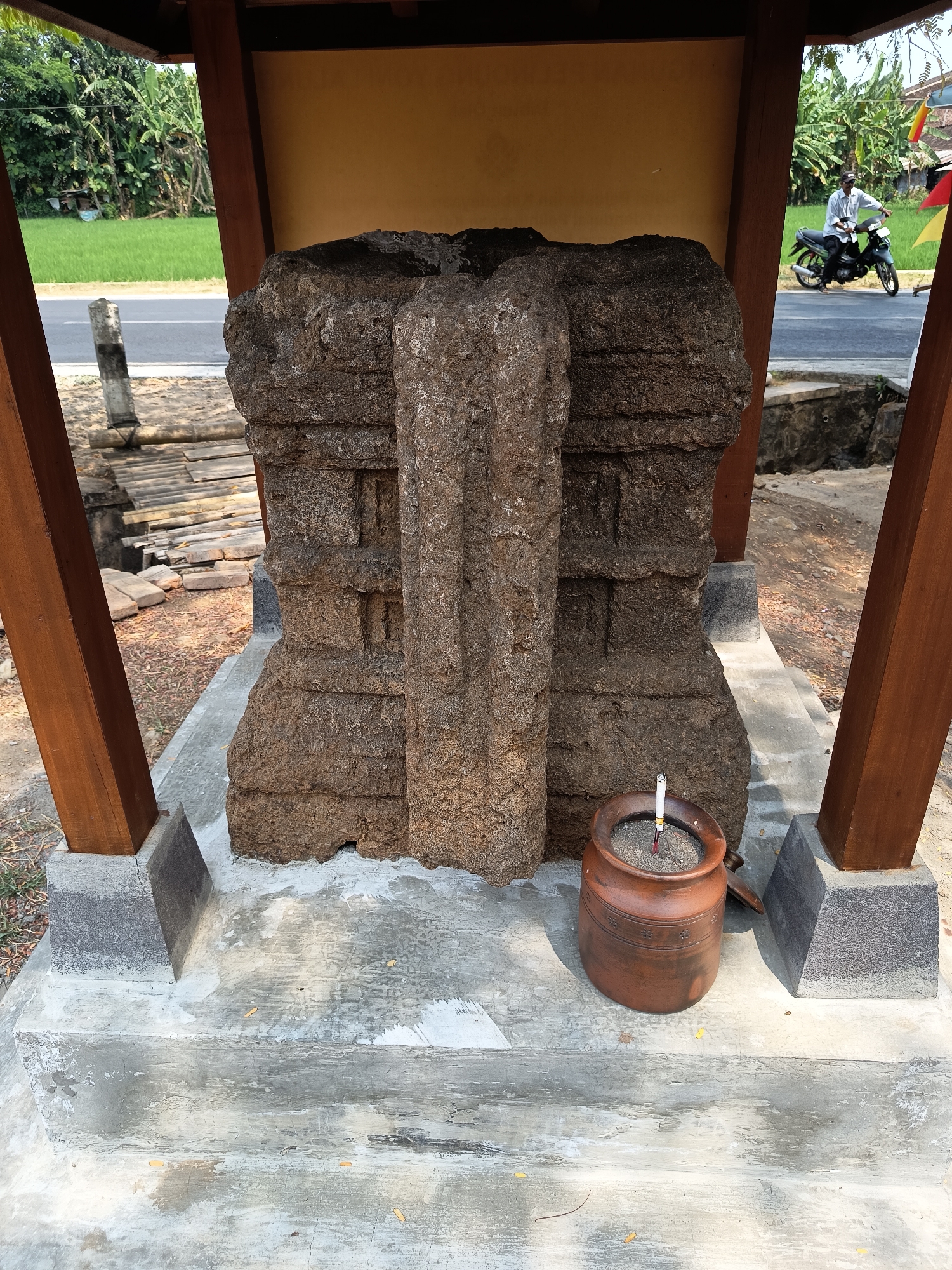 In Lalung, Indonesia, a weathered stone artifact, possibly a historical monument, stands protected by a wooden shelter. The artifact, carved from rough, dark stone, displays layered designs and is positioned on a concrete base. In front of it sits a brown ceramic pot filled with ash and a lit cigarette, suggesting recent use as an ashtray. The background reveals a road with a person riding a motorcycle, lush green fields, and banana trees, indicating a rural or semi-rural environment. Wooden pillars support the shelter's roof, and the overall scene appears to be an outdoor exhibit or monument.