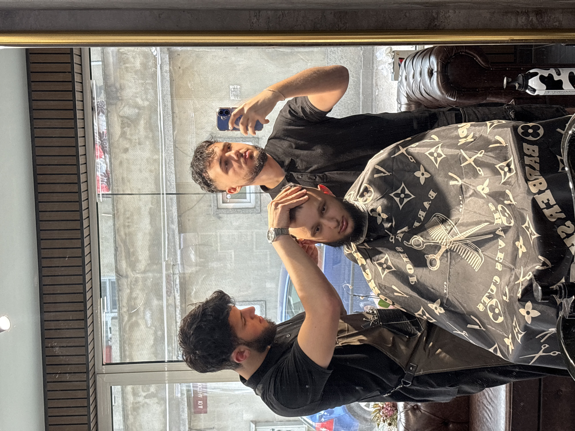 The image presents a barbershop scene in Varna, Bulgaria, captured as a reflection in a large mirror with a gold upper frame. Three men are visible.
In the center, a barber, with dark hair and a beard, wears a black short-sleeved shirt and a dark, possibly leather, apron. He is actively engaged in cutting or styling the hair of a client, his hands positioned near the client's head. A silver watch is on his left wrist. The client, also with dark hair and a beard, sits calmly in a barber's chair, covered by a black barber's cape. The cape features a distinctive pattern of white symbols resembling Louis Vuitton monograms, interspersed with barber tools like scissors and combs, and the reversed text "BYBZR" and "BARBER".
A third man, presumably the photographer, is visible in the upper reflection, holding a blue smartphone to take the picture. He has dark, curly hair and a beard, wearing a black polo shirt, and is looking at the phone screen.
The barbershop interior includes a dark, tufted leather armchair or sofa visible on the right, next to which stands a bottle with a cow print pattern and the word "COW" visible. On the left, a vertical wall panel with horizontal dark wooden slats adds to the modern aesthetic. Through the large window reflected in the background, an urban environment is visible, including a gray building and a partial view of a blue car, illuminated by bright daylight, suggesting a clear, sunny day. Faint reversed text, "ΑΟΙ ΛΗΤΡΟ", is barely discernible on a sign outside. The overall scene depicts a contemporary barbershop in action.