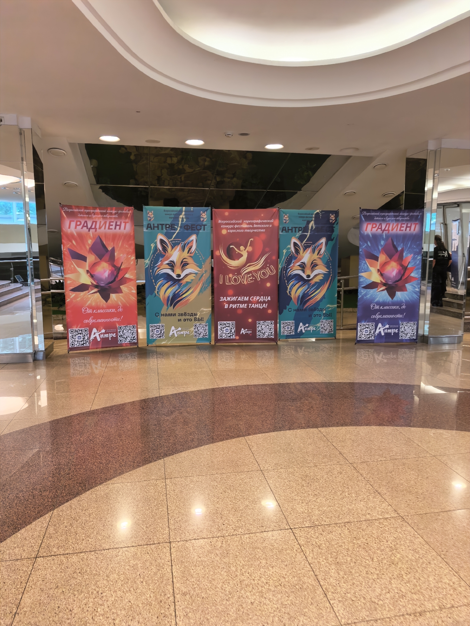 A group of five large, vertical banners are displayed in an indoor location. The banners are spaced evenly and stand upright on a polished floor. Each banner features vibrant graphics and text, suggesting they are for an event or competition.
The leftmost banner is red and features a graphic of a lotus flower with a faceted gemstone at its center. The text "ГРАДИЕНТ" (Gradient) is prominently displayed in large, white letters. Below this, smaller text reads "От классики, до современности!" (From classics to modernity!). At the bottom are two QR codes and a logo.
The second banner from the left is teal and features an illustration of a fox with colorful, flowing lines around its head. The text "АНТРЕ-ФЕОТ" (Entre-Feat) is visible. Below this, it says "С нами звёзды и это Вы!" (With us stars and it's you!). It also includes QR codes and the same logo.
The middle banner is red and features a silhouette of a dancer in a dynamic pose. The text "I LOVE YOU" is in a stylized font. Below this, it reads "ЗАЖИГАЕМ СЕРДЦА В РИТМЕ ТАНЦА!" (We ignite hearts in the rhythm of dance!). This banner also has QR codes and the logo.
The fourth banner from the left is a dark blue with a graphic similar to the leftmost banner, featuring a lotus flower and gemstone. The text "ГРАДИЕНТ" is again displayed in large, white letters, with the same tagline "От классики, до современности!" below. It also has QR codes and the logo.
The rightmost banner is predominantly blue and features a stylized image of a fox, similar to the second banner from the left. The text "АНТРЕ-ФЕОТ" is visible, with the same tagline "С нами звёзды и это Вы!". It also includes QR codes and the logo.
The floor is a reflective, light-colored material with a pattern of darker, reddish-brown granite-like tiles forming a curved section in the foreground. Two bright, circular lights are embedded in the floor, casting reflections. In the background, a person wearing dark clothing is partially visible near a mirrored surface. The overall environment appears to be a modern indoor space, possibly a mall or an event venue.