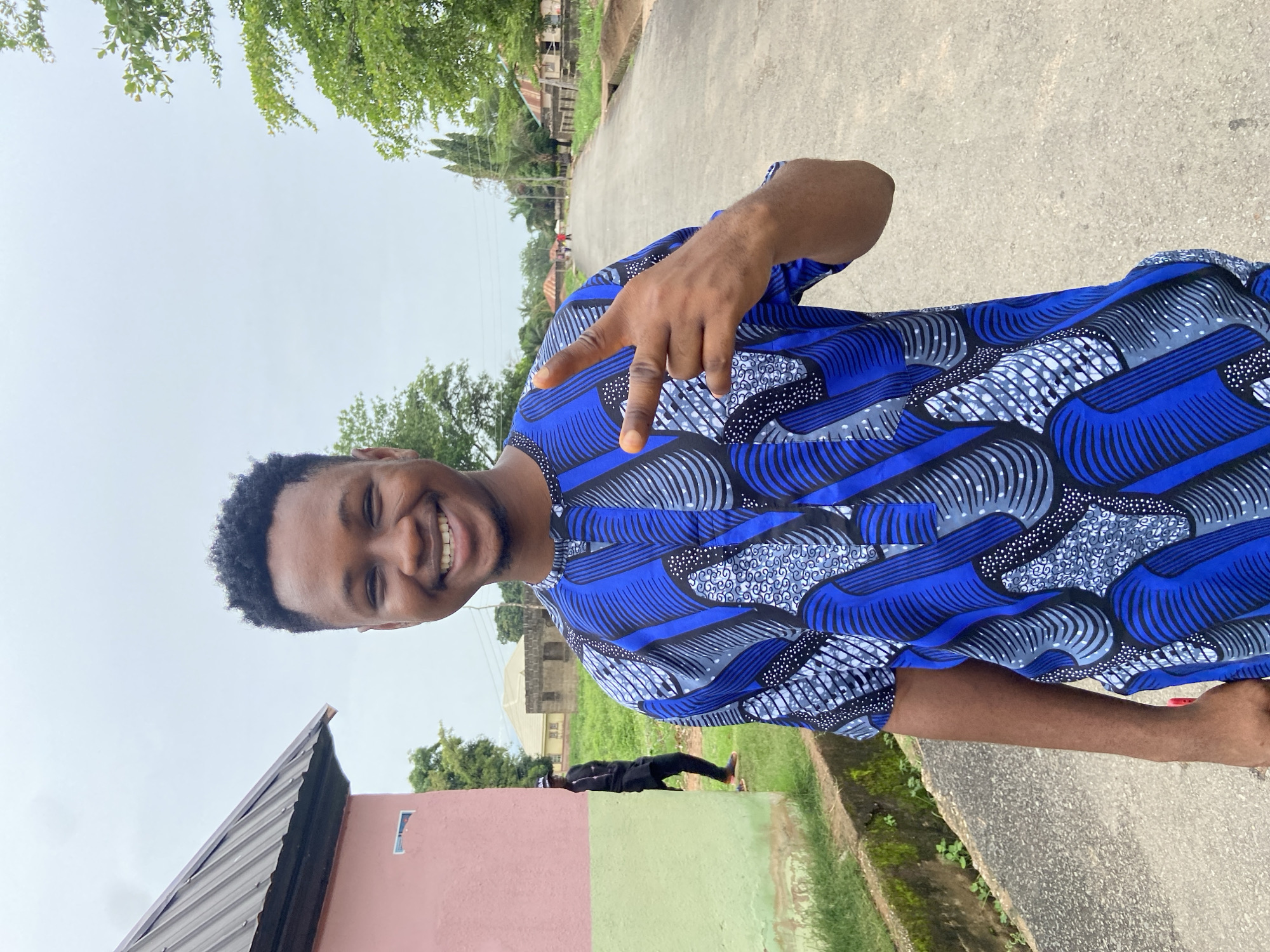 A young man stands on a street in Mararaba, Nigeria. He is wearing a blue patterned shirt and smiling at the camera, while making a peace sign with his right hand. The background includes a road, buildings, trees, and a cloudy sky.