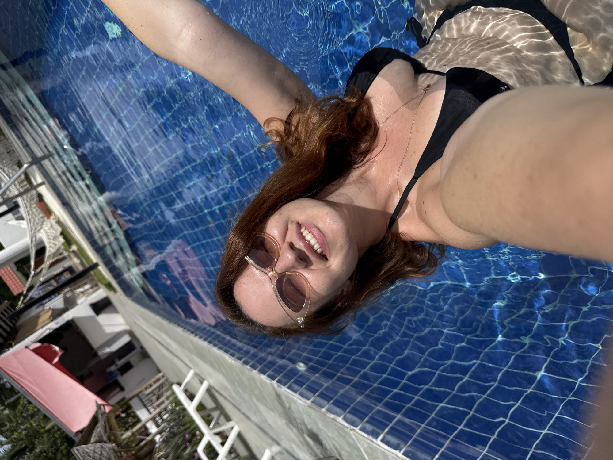 A woman with auburn hair and sunglasses is taking a selfie in a swimming pool in Baan Taling Ngam, Thailand. She is wearing a black bikini top and a gold necklace. The pool water is blue and clear, and the bottom is tiled. In the background, there are lounge chairs and a building with a pink awning. The sun is shining, and the water is reflecting the light.