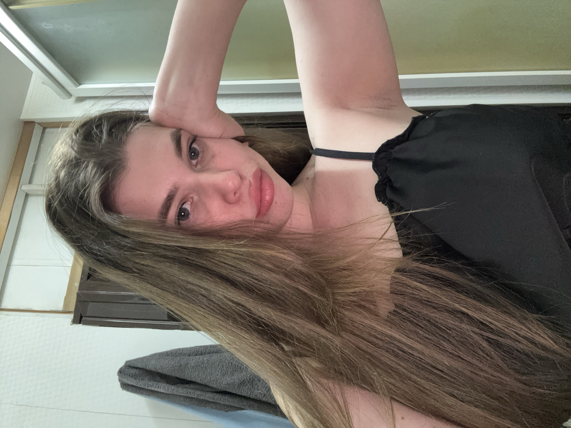 A woman with long, light brown hair is posing for a selfie in an indoor setting, possibly a bathroom. She is wearing a black top with a thin strap and is holding her head with her right hand. Her gaze is directed towards the camera. A towel hangs on the wall, and a window is visible in the background. No location in Мещёры, Russia can be identified in the image.