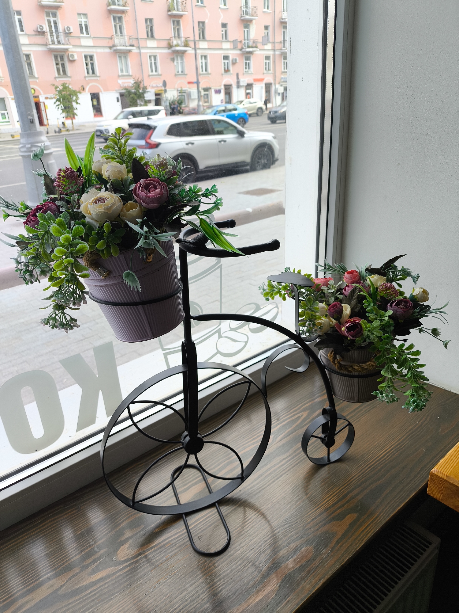 The image depicts two artificial floral arrangements housed in decorative pots mounted on a black metal bicycle-shaped stand. The stand is positioned on a wooden surface next to a large window. Through the window, a street scene in Tver, Russia, is visible, featuring a pink building with balconies, several cars, and pedestrian activity. The floral arrangements consist of various artificial flowers and greenery, with one pot a light purple and the other a muted gray. The overall scene suggests an interior setting with a view of an urban environment.