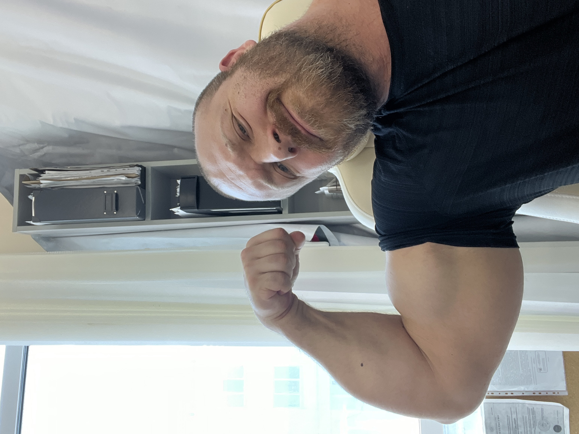 A bearded man with a shaved head is positioned close to the camera, flexing his right bicep. The image appears to be rotated 180 degrees. The man is wearing a black t-shirt and is seated in what appears to be an office chair. Behind him are office shelves containing files and various objects. A window is visible, suggesting a location indoors. Based on the information provided, it is impossible to definitively state this is in the city of Chaikovsky, Russia.