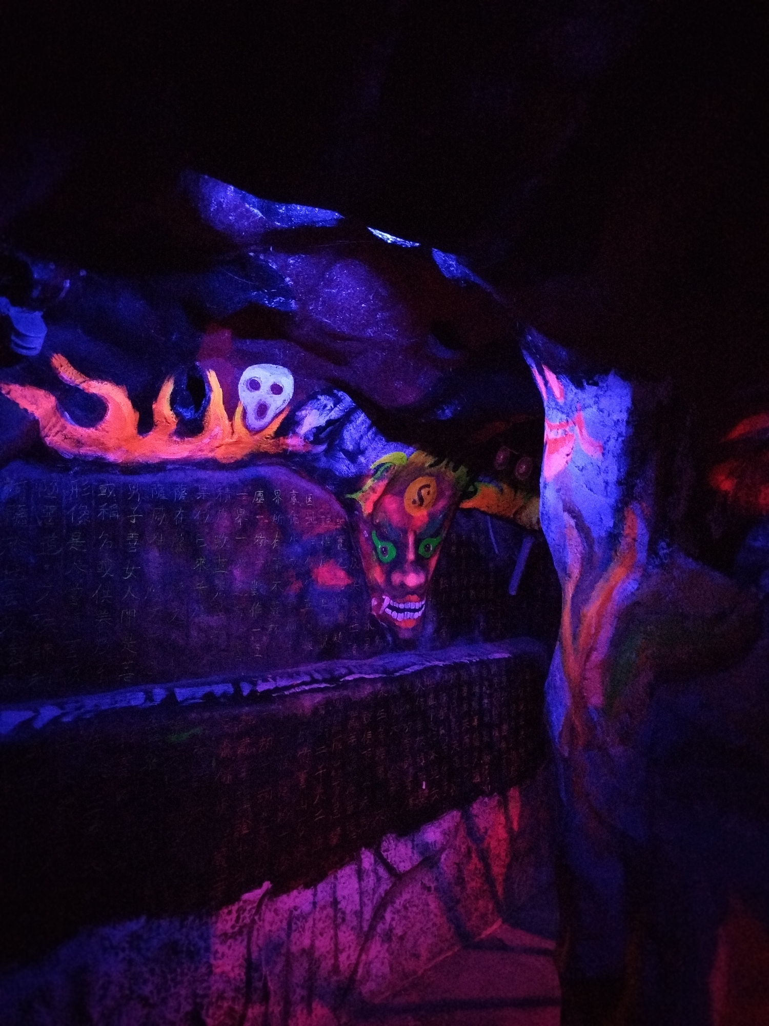 The photograph depicts a dimly lit, possibly artificial cave environment, with artwork on the rock surfaces. On one rock face, there is a series of fluorescent paintings, including a stylized orange flame, a white skull, and a face with green eyes, a red nose, and sharp white teeth. The rock surface also has what appears to be black text written in an Asian language. Another rock formation has fluorescent orange and blue patterns painted on it. The overall lighting is uneven, with strong blue and purple tones enhancing the fluorescent colors, and the setting appears to be enclosed and subterranean. There are no apparent people, subjects, or actions depicted. The location and environment are a decorated cave-like space. Unknown city and Unknown location.