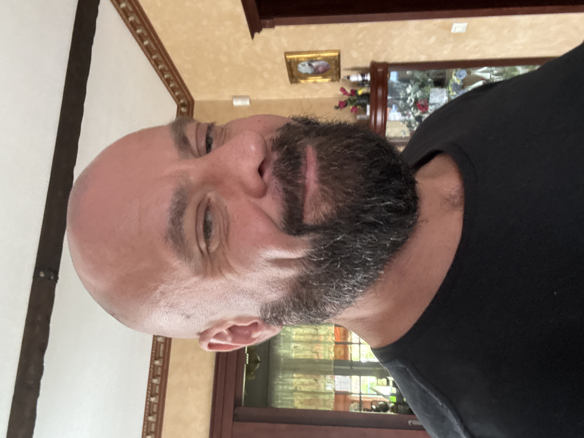 A man with a shaved head and a beard is looking at the camera and smiling. He is wearing a black t-shirt. The background is a room with light yellow walls and dark wooden trim. There is a framed picture on the wall and a display cabinet with flowers in it. The image appears to be taken in a residential setting.
