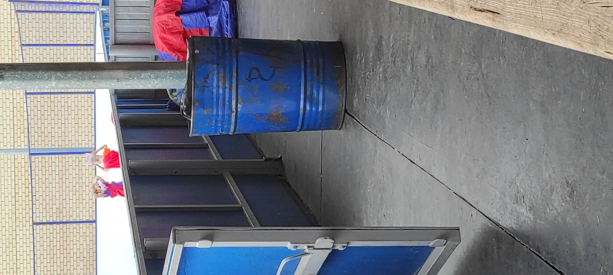 The image appears to depict an outdoor scene, possibly an industrial or recreational area, given the presence of a blue barrel, a wooden structure, and what looks like a fence with a brick wall in the background. Two young children are visible in the distance, standing behind a railing. They are wearing brightly colored clothing, one in purple and the other in orange. They appear to be holding up some papers or objects. The ground is paved with concrete, and there are some blue structures on the left side of the image. The overall lighting suggests it is daytime. The location is specified as Noviy, Russia.