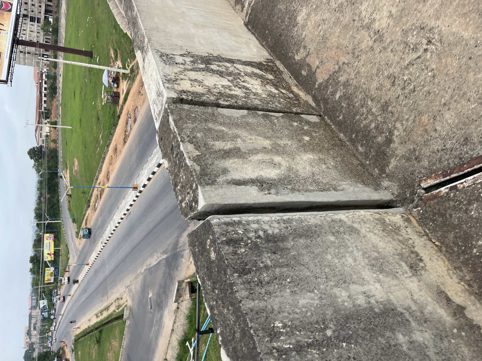 The image shows an elevated view of a section of road in Osogbo, Nigeria. The camera is positioned above a concrete barrier, with the road stretching into the distance. A median with alternating black and white stripes divides the road. Vehicles, including cars and motorcycles, are visible on the road. Buildings, billboards, and green spaces line the road. A small market area is visible to the right of the road in the middle distance. The sky appears overcast. The concrete barrier in the foreground is stained and shows signs of weathering.
