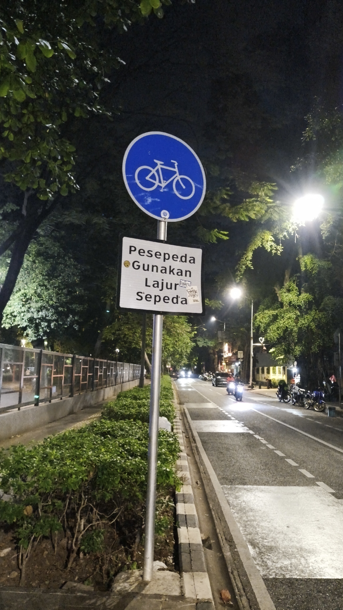 The image shows a street scene at night in South Jakarta, Indonesia. A blue circular sign with a white bicycle symbol is mounted on a pole. Below it, a white rectangular sign reads "Pesepeda Gunakan Lajur Sepeda", which translates to "Cyclists Use Bicycle Lane". The sign has graffiti on its lower right corner. The street has a designated bicycle lane marked by white dashed lines and rectangular white areas. Traffic, including cars and motorcycles, is visible on the road. Trees and bushes line the sidewalk, and streetlights illuminate the scene. A metal fence runs along the left side of the sidewalk.