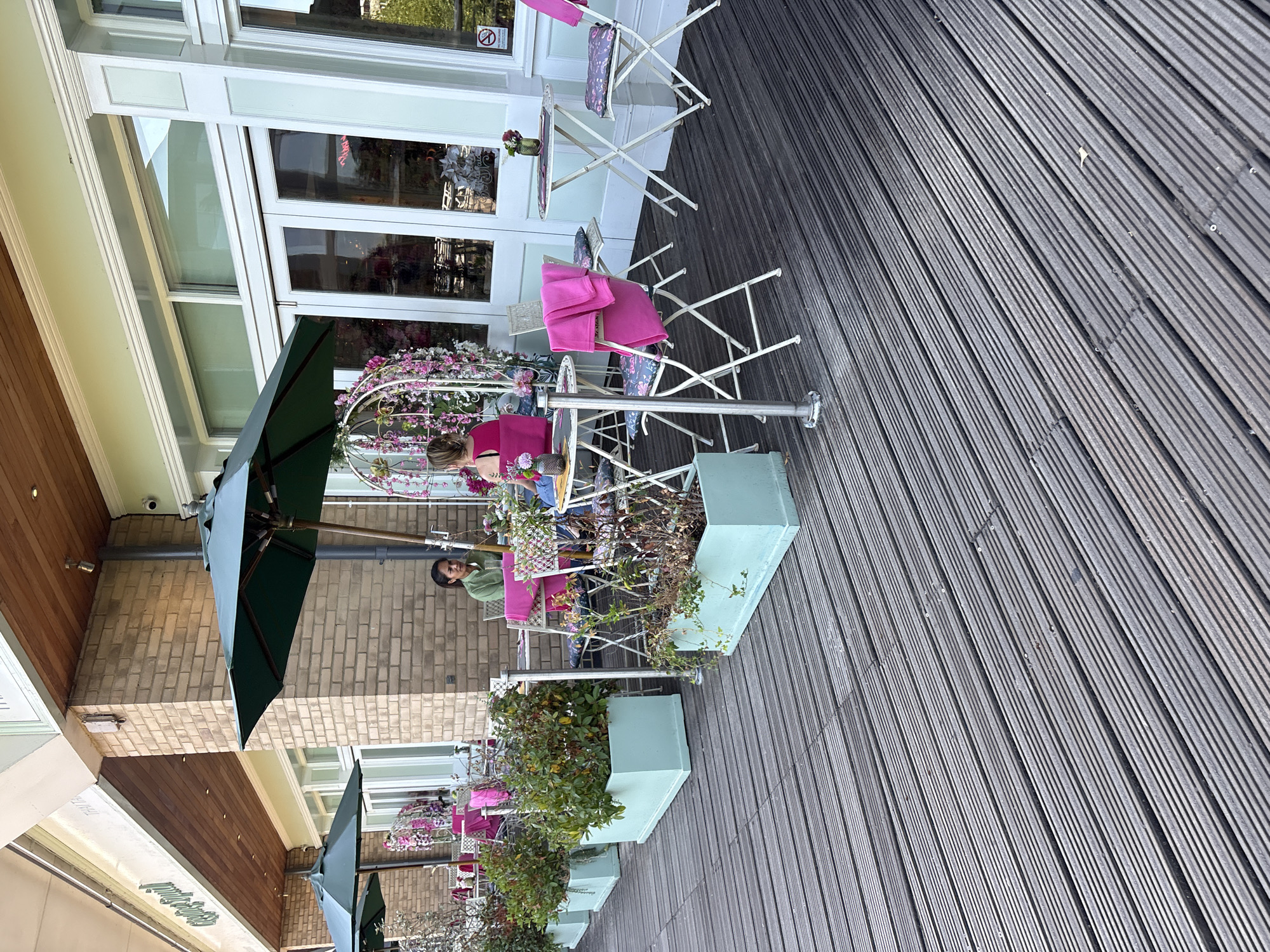 The image shows the outdoor seating area of a restaurant, likely located in Chelmsford, United Kingdom, given the signage visible which includes "Sterling" and "Thai". The seating area features a dark wooden deck with multiple small, round tables and white, foldable chairs. Some tables are adorned with floral arrangements and a pink blanket is draped over one chair. Two women are seated at separate tables. One woman, wearing a green top, is seated at a table with a pink cushion. The other woman, wearing a pink top, is seated at a table adorned with a white decorative birdcage filled with pink and white flowers. In the background, the restaurant has large glass doors and windows, with a brick facade and a wooden overhang. Several large, light blue planters containing green foliage are positioned along the edge of the deck, separating it from the restaurant's facade. Green umbrellas are also present, providing shade for the seating area. The overall scene suggests a pleasant outdoor dining experience.