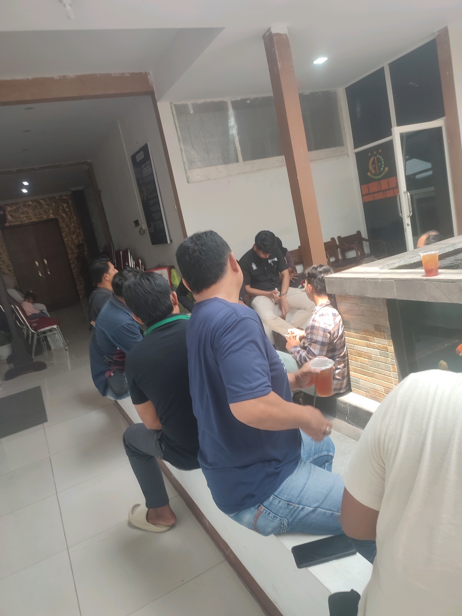 In a well-lit interior space, likely a waiting area or common room in Nganjuk, Indonesia, a group of men are seated. The environment features tiled flooring and light-colored walls, with wooden beams supporting the ceiling. The scene is divided by a low, stone-tiled partition, possibly a decorative element or a planter box.
At the far end of the room, a woman is seated on a chair in what appears to be a separate, more private area. To her left, a large, ornate wooden door is visible.
In the foreground and midground, several men are interacting or observing. One man, wearing a black polo shirt, is seated and appears to be engaged in conversation. Another man, in a plaid shirt, is holding a cup of amber liquid, possibly tea or iced coffee, and is also seated. A man in a dark blue t-shirt is in the process of holding a cup, while another man, wearing a black t-shirt with a green lanyard, is seated. In the immediate foreground, the back of a man wearing a light-colored t-shirt is visible, with a smartphone lying on a white ledge in front of him. The overall atmosphere suggests a casual gathering or waiting period.