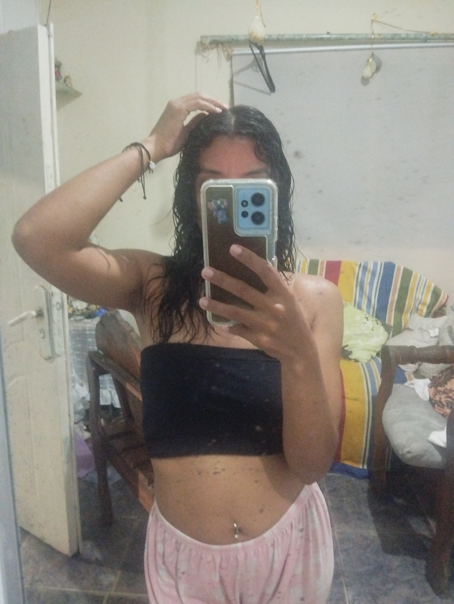 A person stands in front of a mirror in what appears to be a domestic setting. The individual is wearing a black strapless top and pink patterned pajama bottoms, and has a navel piercing. Their hair is dark and wet, with one hand touching their head. The person is holding a smartphone, presumably taking a selfie. The reflection in the mirror shows a partially visible room with furniture, including chairs, and a striped cushion. The background also reveals a door and a wall with some items hanging. The overall lighting is subdued. There is no information available in the image to identify the location as El Valle del Espíritu Santo, Venezuela.