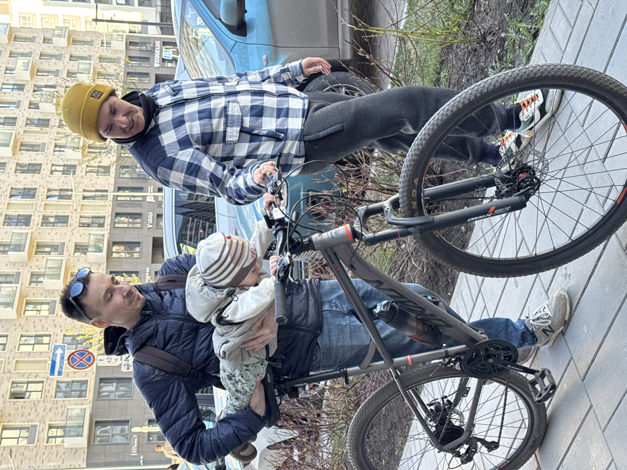 Two adult males are observed interacting with a child and a bicycle in an urban setting. One adult, wearing a yellow beanie and a blue-and-white plaid jacket over dark pants, is positioned on the right side of the frame. The second adult, wearing a dark-colored jacket and sunglasses atop his head, holds a child in a white jacket and patterned pants on the left side of the frame. The child is facing towards the handlebars of a gray mountain bicycle. The bicycle's rear wheel is on the sidewalk. In the background is a blue car and a building with repeating window patterns. A blue and white "no stopping" street sign is visible on the left side of the frame. The location is identified as Izhevsk, Russia.