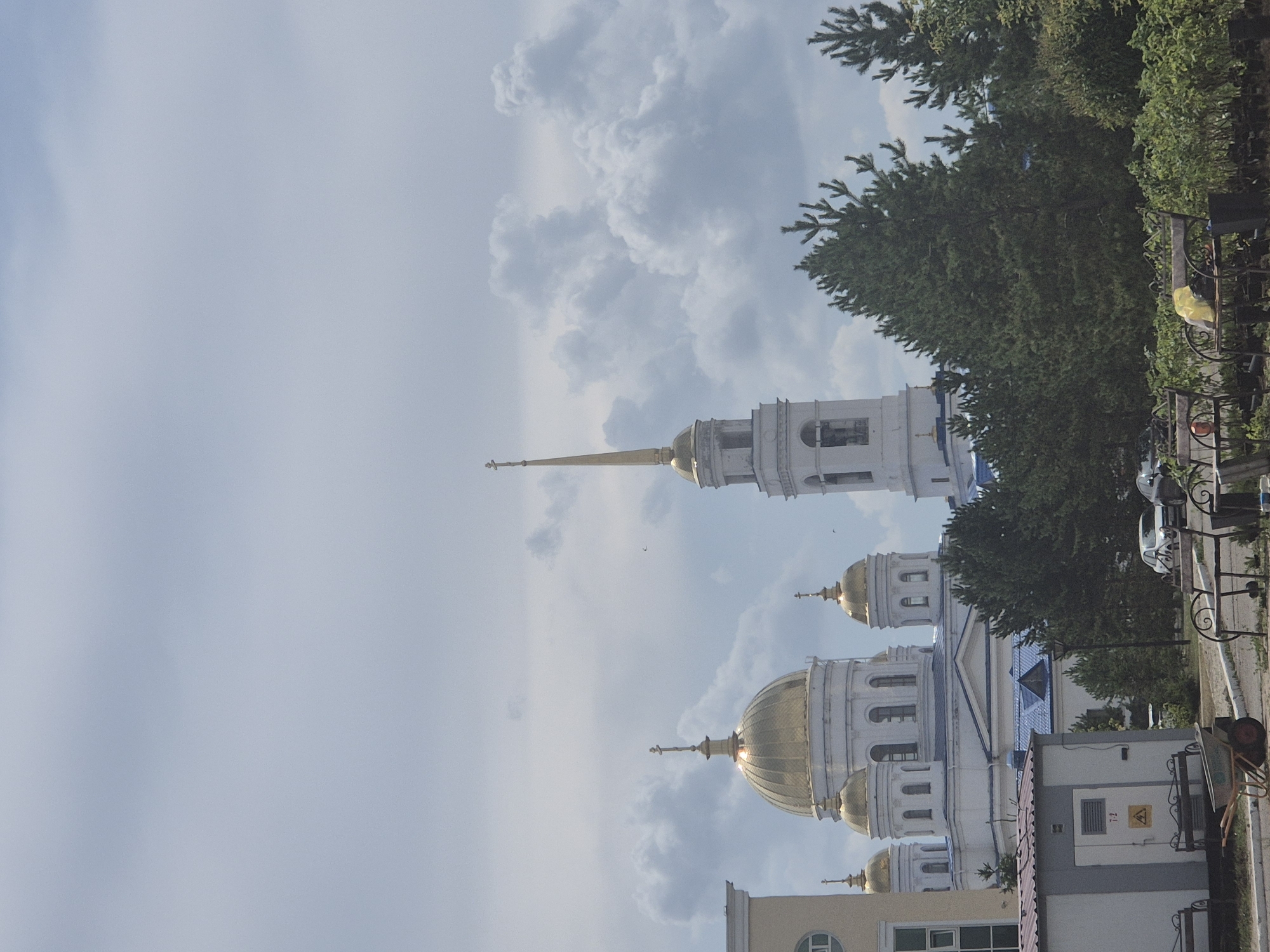 The image shows a view of the Ascension Cathedral in Kokshetau, Kazakhstan. The cathedral features prominent golden domes and a tall white bell tower. In the foreground, there is a small utility building with electrical warning signs, and several green trees. There are also some park benches and a wheelbarrow in the foreground. The sky is cloudy.