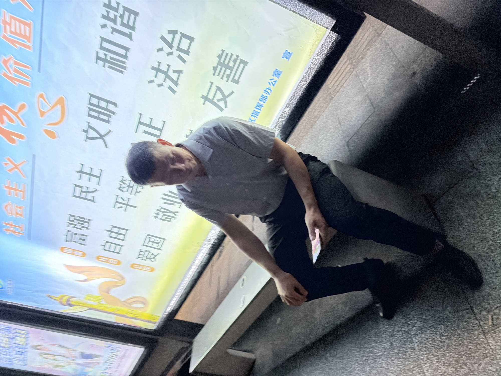 A man sits on a bench in front of a brightly lit billboard in Jiangjin, China. The billboard features Chinese characters, including "社会主义核心价值观" (Socialist Core Values). The man, wearing a light-colored short-sleeved shirt and dark pants, holds a smartphone in his right hand. The background shows the illuminated advertisement and a portion of another display to the left. The foreground consists of a textured pavement and shadows. The overall scene appears to be an outdoor public space at dusk or night.