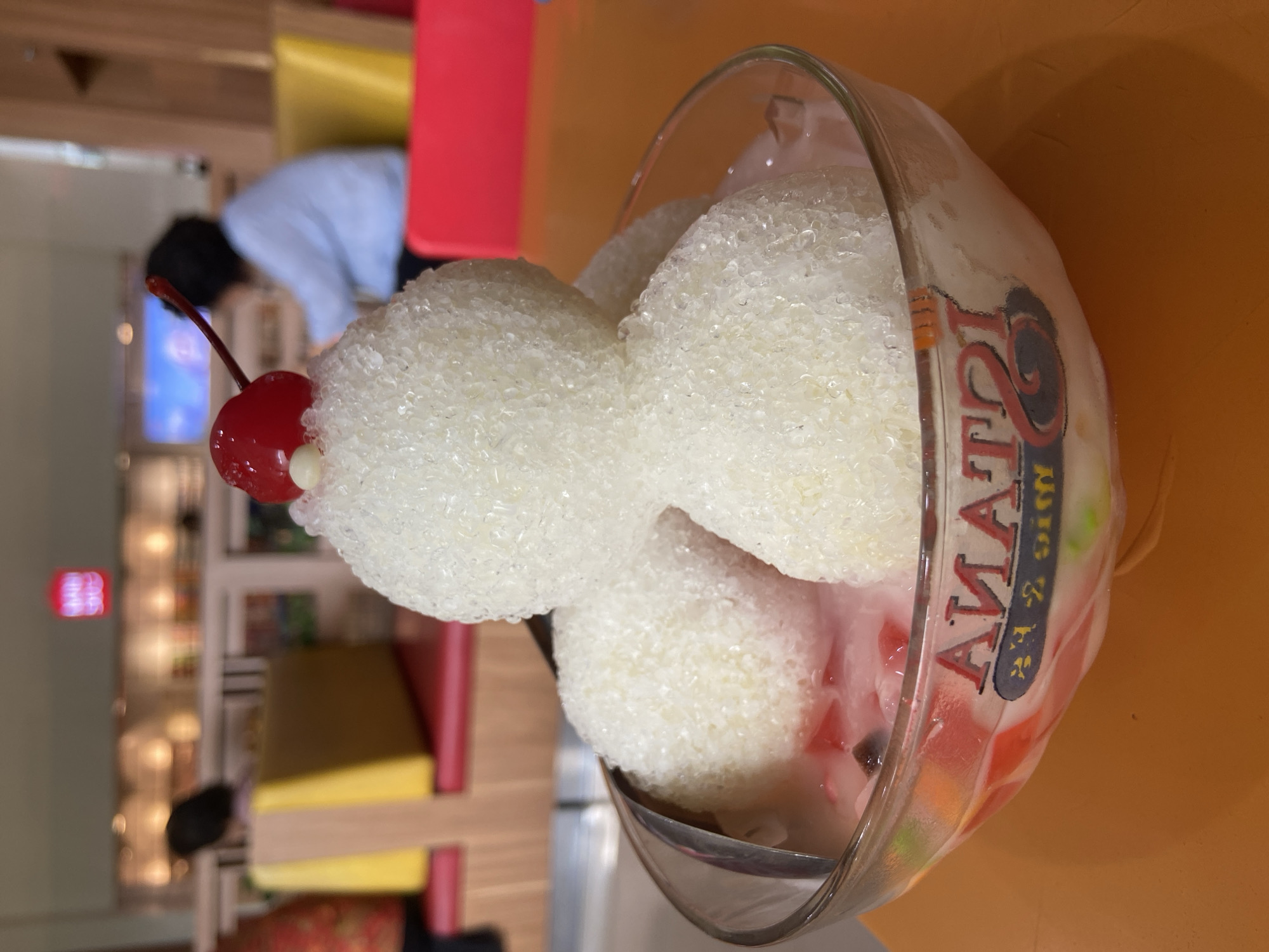 A close-up, eye-level shot features a dessert in a clear glass bowl filled with a pink liquid and colorful fruit pieces. The dessert itself consists of four scoops of white, granular ice, with a maraschino cherry and a white bead topping the central, uppermost scoop. A silver spoon is partially visible on the left side of the bowl. The bowl rests on an orange table. In the blurred background, several people are seated at tables in a restaurant setting. One person in the mid-ground wears a light blue shirt, and another person in the foreground-left is partially visible wearing a patterned orange shirt. Yellow and red seating are also visible in the background. The restaurant appears to be "TATA" based on the text visible on the bowl. The overall scene suggests a dessert being served in a casual dining establishment.