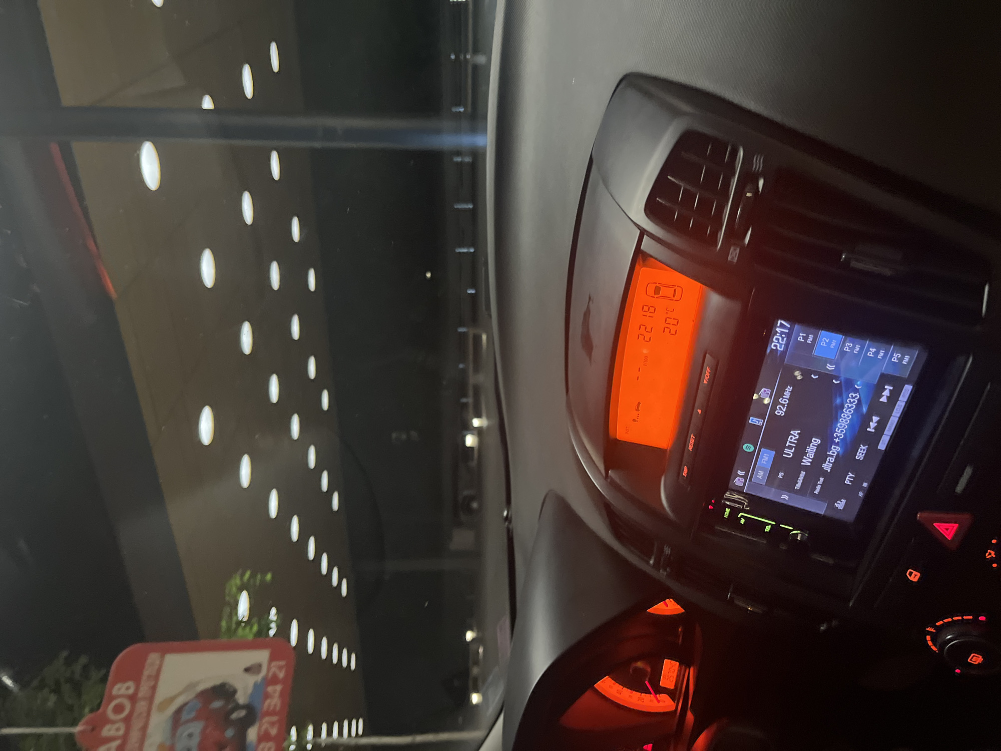 The image displays the interior of a car at night, looking out through the front windshield. The dashboard occupies the right side of the frame, illuminated with orange and blue lights from the car's displays. The main display shows the time as "22:18" and an interior temperature of "20°C." Adjacent to it, a car stereo/infotainment screen indicates the time as "22:17" and the radio station "ULTRA" playing on "92.6 MHz." Visible text on the radio screen also includes "ultra.bg" and a phone number, "+359886333." Air vents, control knobs, and a hazard light button are also part of the dashboard.
Outside the car, or reflected on the windshield, is a large, dark building featuring a distinct pattern of numerous circular, brightly illuminated windows or lights. A red sign, partially visible in the bottom-left, contains the text "ABOB" and numbers "32134 21." The overall setting is dark, confirming the late evening or night time indicated by the car's clock. The scene is likely set in Blagoevgrad, Bulgaria, with the vehicle appearing to be stationary or moving slowly.
