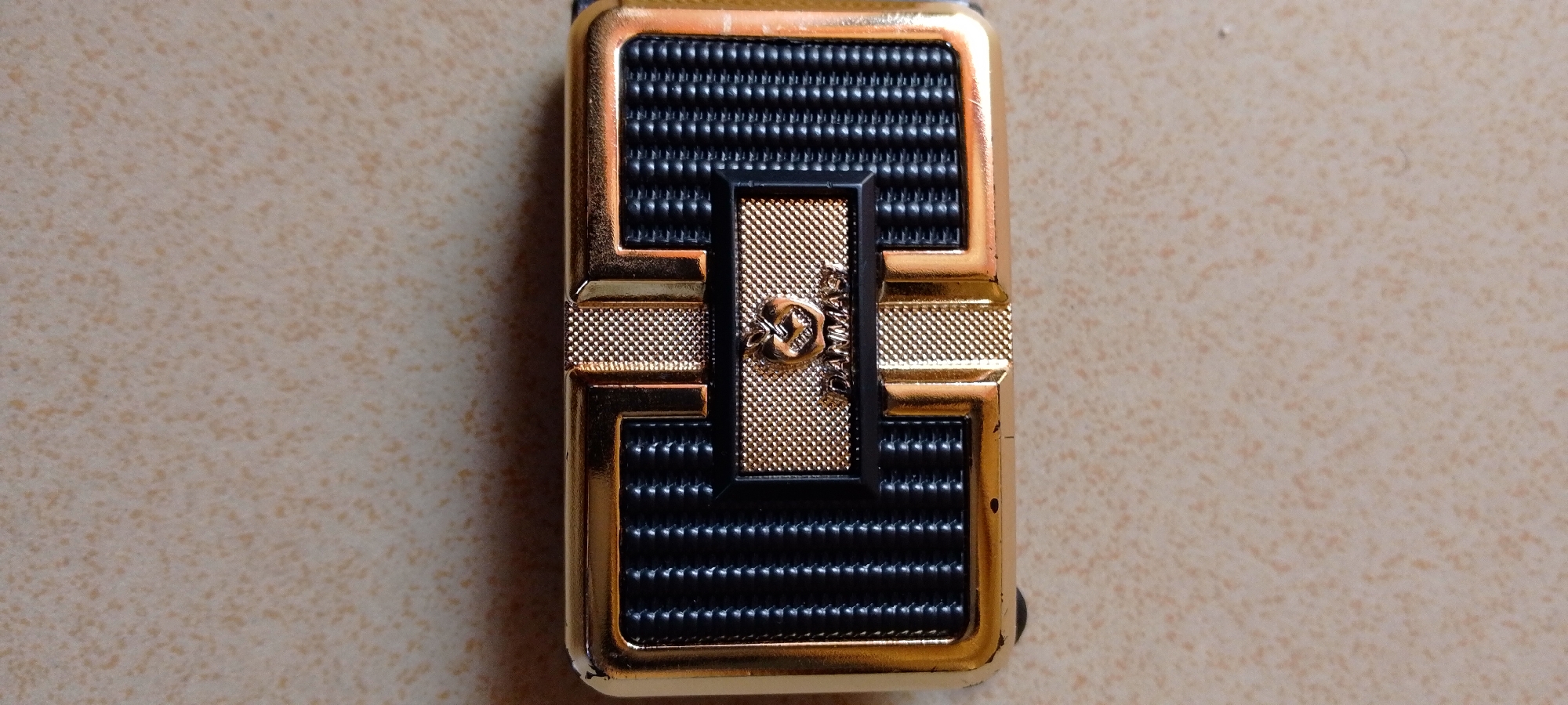 The image shows a close-up, top-down view of a golden and black lighter resting on a textured, light brown surface. The lighter is rectangular with rounded corners and features a decorative pattern. The top and bottom sections of the lighter are covered in parallel black ridges, while the middle section has a textured gold design with a smaller black inset. A small, circular emblem with what appears to be a lion or mythological creature is visible on the gold inset. The surface the lighter is on has a speckled appearance, with irregular patches of darker brown on a lighter tan background. The overall lighting is even, with no harsh shadows. There is no indication of the city of Jalingo, Nigeria, in the image.