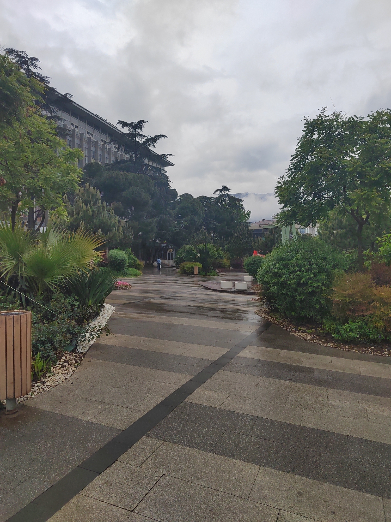 A paved pedestrian area with gray and dark gray stripes stretches into the distance, flanked by vegetation and trees. On the left, there's a wooden bin and dense greenery, while the right features shrubs and trees. In the background, a large building is visible, partially obscured by the trees, and a person with an umbrella walks away on the path. The sky is overcast, suggesting a wet or rainy day, and the pavement appears damp, reflecting the ambient light. Some mountains are partially visible in the background.