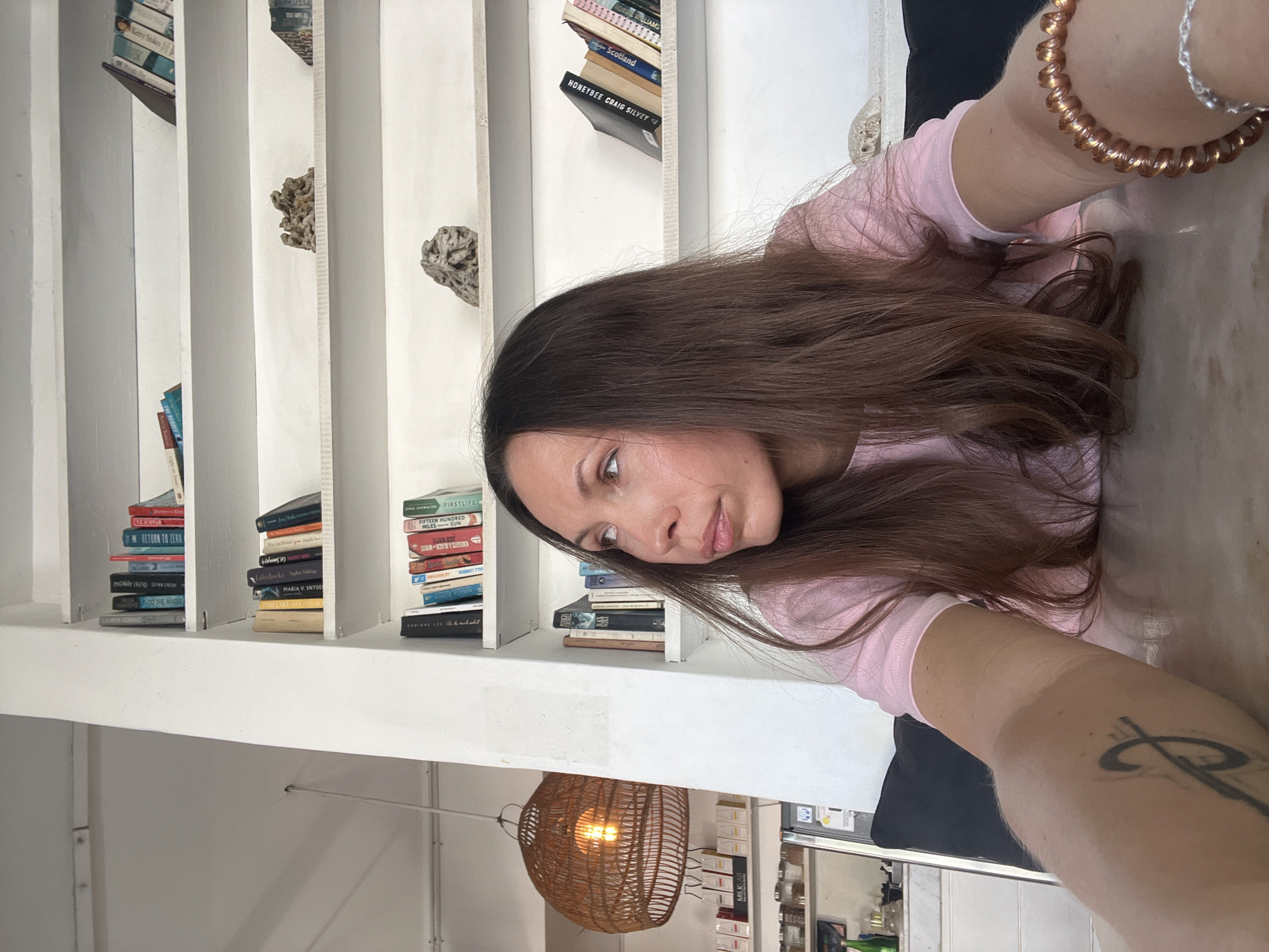 The image is a selfie taken by a woman in what appears to be a room with built-in bookshelves. The bookshelves are white and contain books and decorative stones. Books visible in the background include "VE SCHWAR," "RETURN TO ZERO," "INTO THE WATER," "Lifeshocks," "MARIA V. SNYDE," "FIRSTLIFE," "FIFTEEN HUNDRED SUN," "KNIGHTS A BLACK-WHITE," "Scotland," and "HONEYBEE CRAIG SILVEY." The woman is wearing a pink shirt and has dark hair. She has bracelets on her wrist. A tattoo is visible on her arm. A woven light fixture hangs from the ceiling. A glimpse of a kitchen or service area with shelves of packaged goods (labeled "MILKLAB") is visible in the background. While no specific landmark indicates North Kuta, Indonesia, the image is consistent with the interior design and retail offerings found in that area.