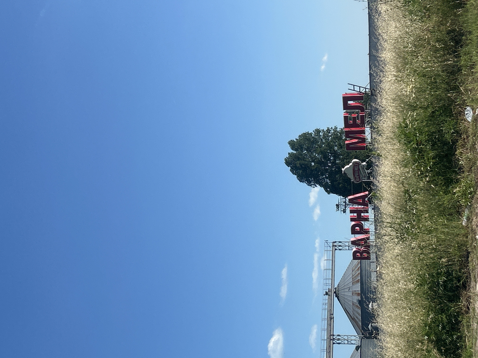 This is a photograph taken in Varna, Bulgaria, featuring a sign for "VARNA MEL" (ВАРНА МЕЛ) which translates to "Varna Mill". The sign is composed of large, red, illuminated letters mounted on top of a building. To the left of the sign are industrial silos, typical of a grain processing facility. The foreground is dominated by tall, dry grass, suggesting an outdoor, possibly rural or industrial edge-of-town setting. The sky above is a clear, bright blue with a few scattered white clouds. The overall scene depicts a working industrial facility against a backdrop of a clear sky.