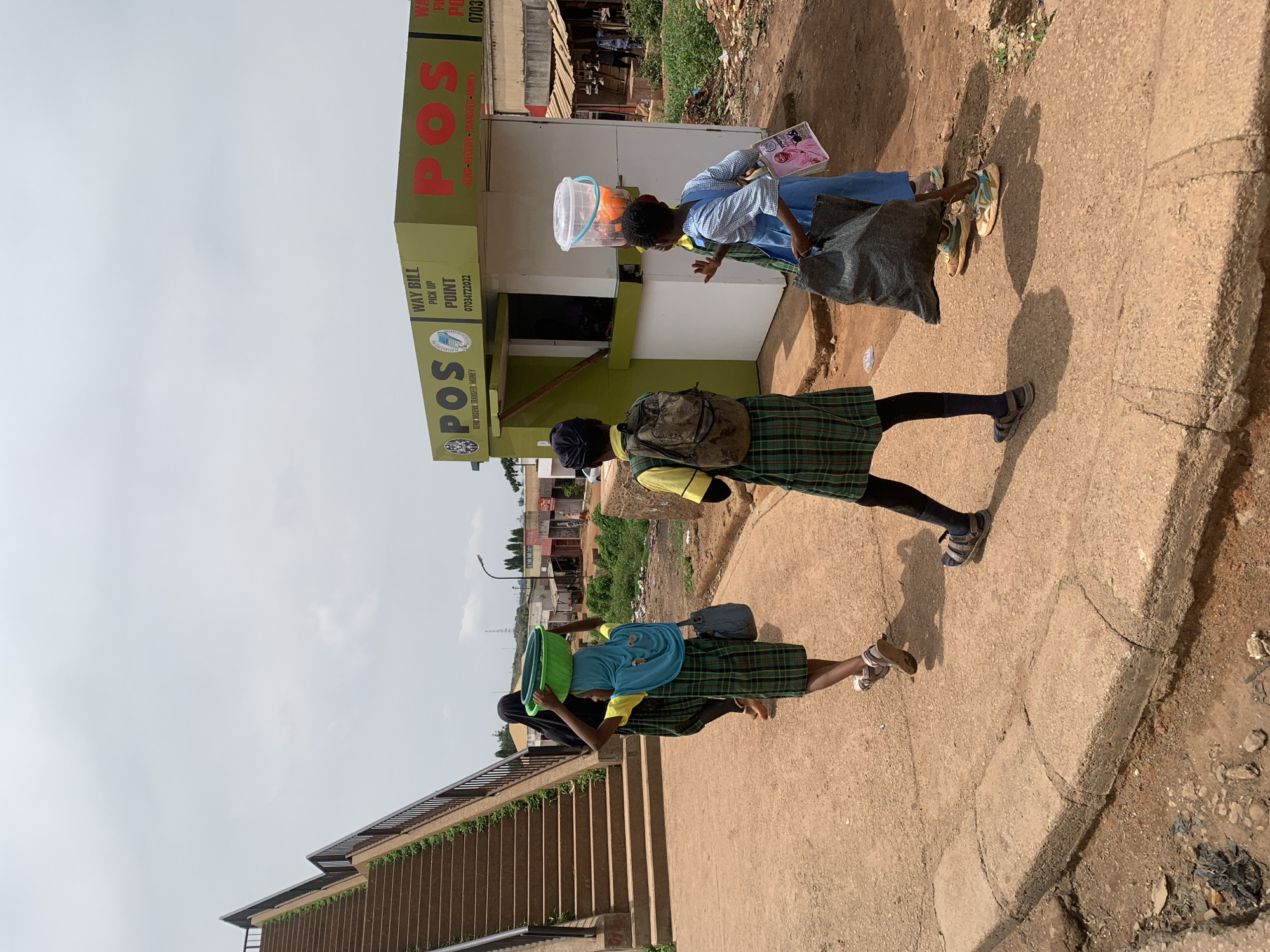 In Gura Top, Nigeria, three young girls are walking on a dirt path alongside a concrete walkway. Two of the girls are wearing school uniforms, consisting of plaid green skirts and yellow tops. One of them has a backpack on. The third girl, in front, is wearing a blue dress and carrying a large black bag in her left arm. The girl in the middle is wearing a blue headscarf and carrying a green basin on her head. She is also wearing a plaid skirt and a yellow top. To the right of the girls is a small green and white kiosk with "POS" prominently displayed on its side, indicating it is a point of sale or money transfer service. A clear plastic container filled with orange and blue items is balanced on the head of another girl, who is partially visible to the right of the kiosk. In the background, there are buildings, some appearing to be shops or homes, and vegetation. To the left, a concrete staircase with dark metal railings ascends upwards. The sky is overcast.