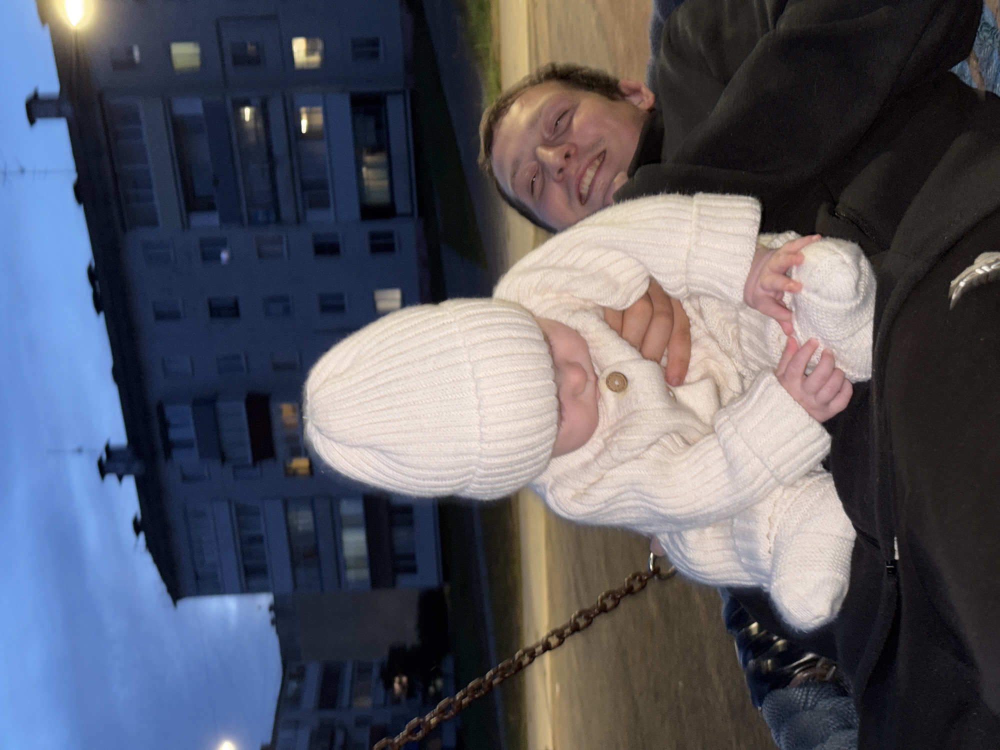 In Noviy, Russia, at dusk, a man holds a baby on a swing. The baby, dressed in a white knitted outfit and hat, is positioned in front of the man. The man, partially obscured by the baby, smiles broadly, his eyes closed. Behind them, apartment buildings with illuminated windows are visible under a dusky sky. A swing chain hangs to the left, and the swing seat appears to be covered in dark fabric with a blue patterned blanket.
