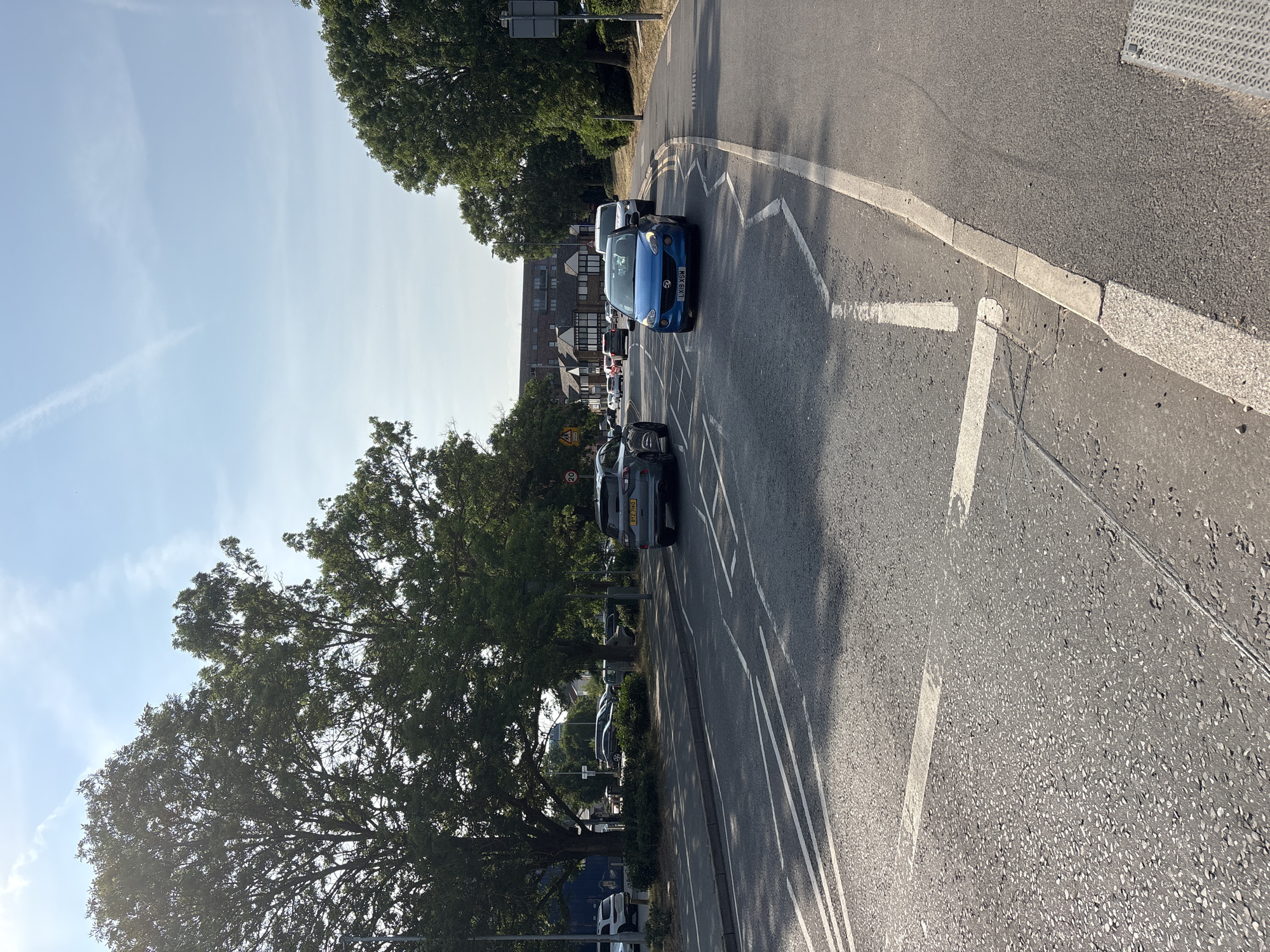 This is a slightly tilted, eye-level, outdoor shot of a residential street in Chelmsford, United Kingdom. The scene is bathed in bright, natural sunlight, suggesting it's daytime, likely late morning or early afternoon, with a clear blue sky and a few wispy clouds. The weather appears to be warm and pleasant.
The primary focus of the image is the road, which is asphalt and has white line markings for a parking bay and a hatched area. Several cars are parked on the side of the road and in the parking bay. The most prominent car is a bright blue hatchback with the license plate "LX61 GXM". Behind it, a dark grey SUV with license plate "BZ17 MMJ" is visible. Further down the street, other vehicles are parked, and there appears to be a residential building with several floors in the background, suggesting a suburban or urban environment.
Lush green trees line the left side of the road, casting dappled shadows on the pavement and road. One large tree in the foreground is particularly dense with foliage. There are also some smaller bushes and hedges visible. In the distance, more buildings and trees can be seen. A yellow triangular road sign with a black border and a pedestrian symbol is visible on the left side of the road, indicating a pedestrian crossing. Another sign with "20" is visible, likely a speed limit sign.
The overall impression is one of a quiet, sunny day in a residential area. There are no people clearly visible in the frame. The scene is static, with cars parked and no immediate activity taking place. The light suggests a clear day, and the presence of mature trees and residential buildings firmly places the scene in a developed area.
