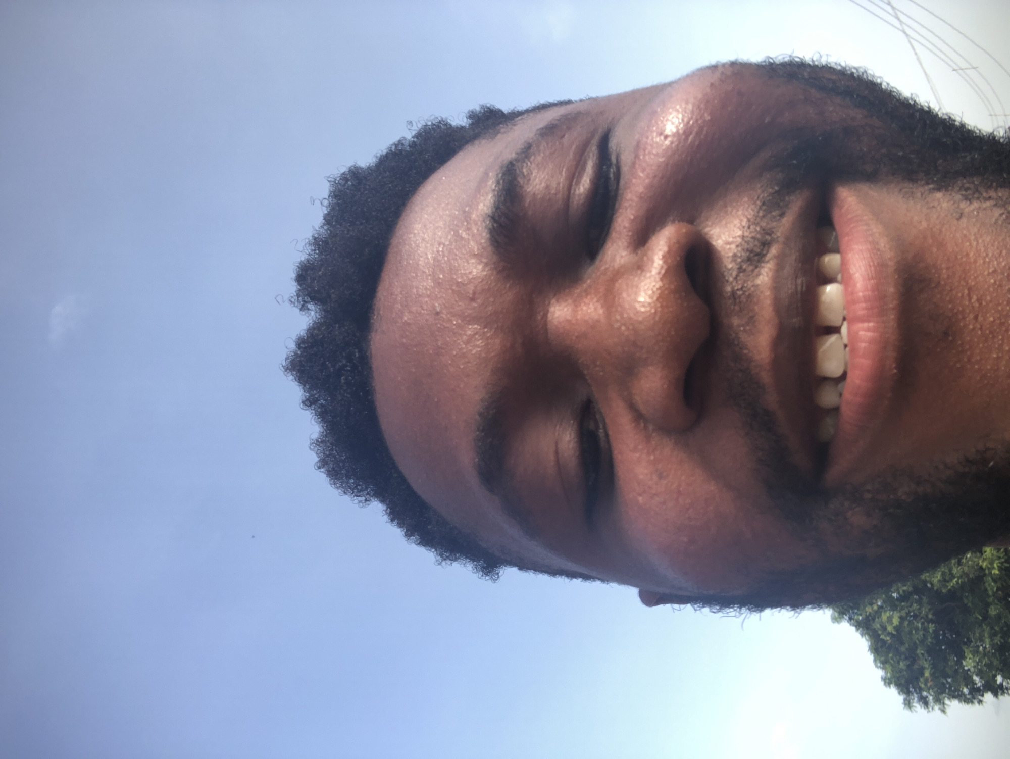 A close-up, low-angle shot shows a man's face against a bright, blue sky. The man has dark skin, short, curly black hair, and a beard. He is smiling, revealing his teeth. His eyes are partially closed. The man's forehead and face are visibly moist, possibly from sweat. In the lower right corner, a portion of a green tree or foliage is visible.