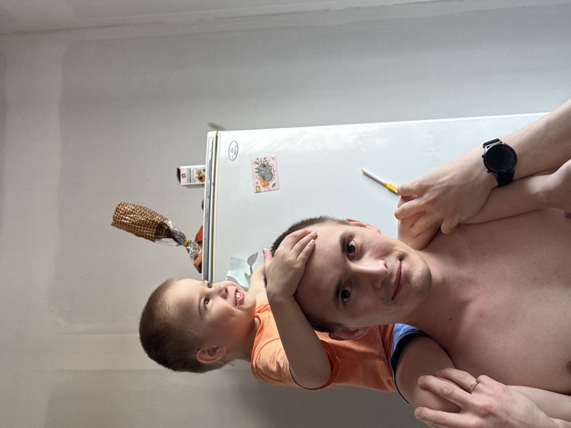 The original image, presented sideways, captures a joyful indoor scene, likely in a casual home in Chaikovsky, Russia, featuring a man and a young child. The man, possibly the father, is seen shirtless with short dark hair, looking directly at the camera with a gentle smile. He wears a black smartwatch on his left wrist and a ring on his right hand. A young child, likely a boy, with light brown hair and wearing an orange t-shirt, is positioned above the man, possibly on his shoulders or being held aloft. The child is smiling broadly and looking upwards and to the left, with his hands playfully placed on the man's head, indicating a happy and affectionate interaction.
In the background, a white refrigerator is visible, decorated with several magnets, one notably featuring a gray creature holding a plant. A yellow and white pen or similar object rests on top of the refrigerator near the man's arm. A crumpled brown bag, possibly containing snacks, hangs from the top edge of the refrigerator on the left side of the frame. The walls are a plain, light color, suggesting an unadorned or in-progress domestic setting. No text is clearly legible in the image.