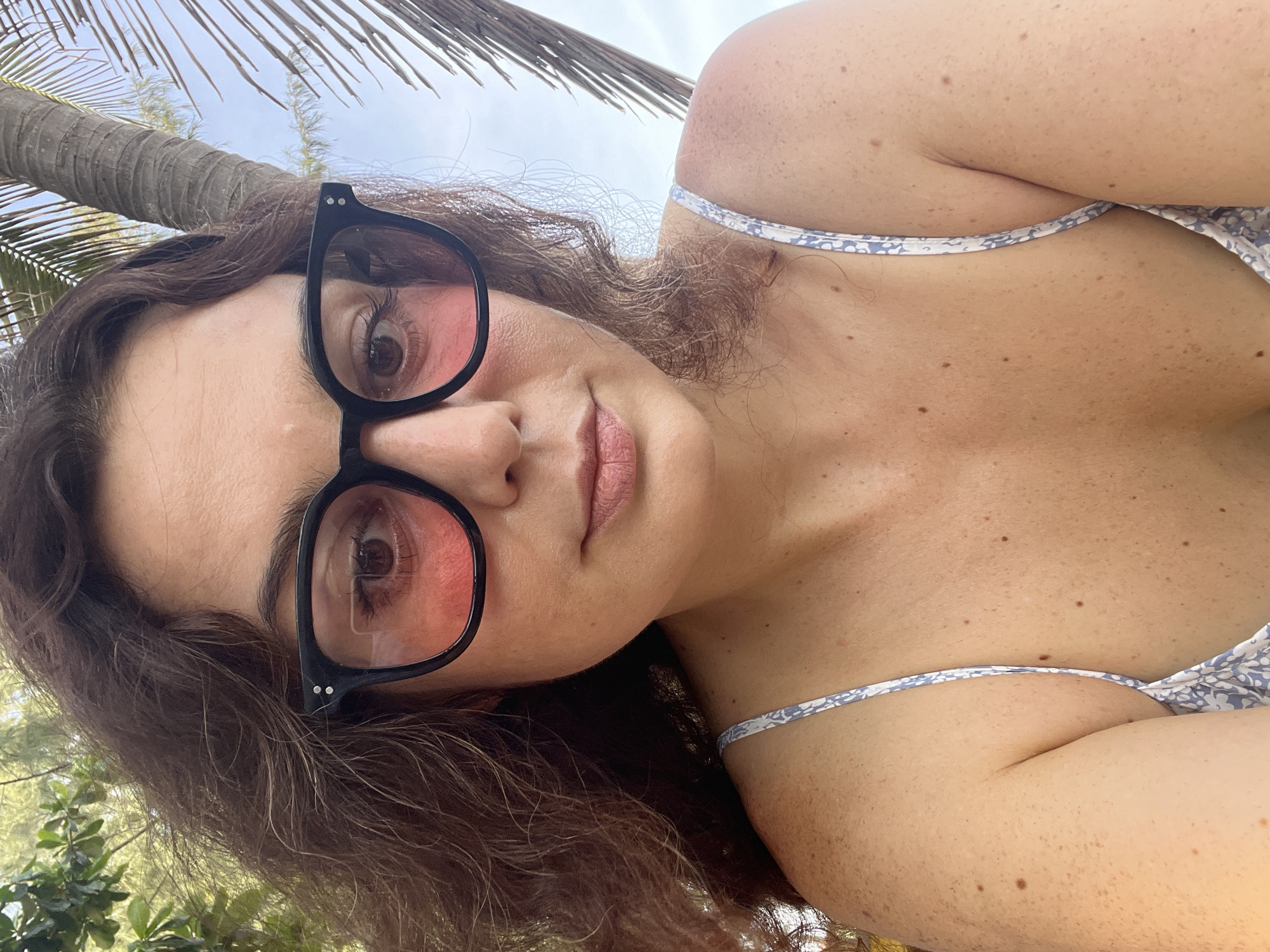 The image is a close-up, selfie-style photograph of a woman with curly brown hair and fair skin, wearing large black-rimmed glasses with pink lenses. She is looking directly at the camera. She is wearing a light-colored camisole with a blue floral pattern. The background shows foliage and a palm tree, suggesting an outdoor, possibly tropical, setting. The overall lighting suggests it is daytime. The image does not provide any information about the location in Phu Thuy Ward, Vietnam.