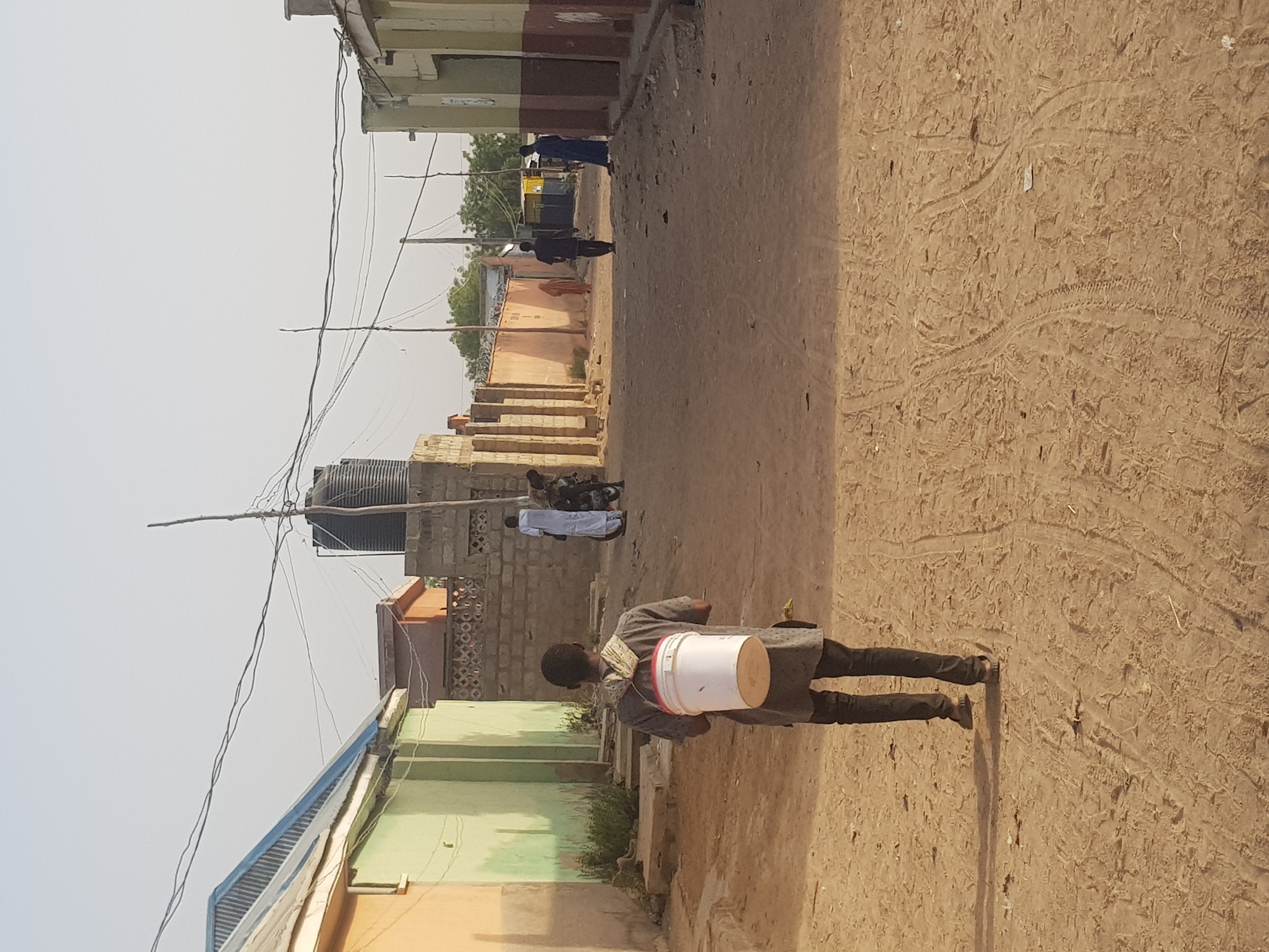 The image captures a street scene in Dabawa, Nigeria. The street is unpaved, revealing sandy, dirt ground with tire tracks visible. A person is walking away from the viewer, carrying a white bucket with red trim over their shoulder. In the background, several people are visible further down the street. Buildings line both sides of the street, constructed from a mix of concrete and brick. A large, black water tank sits atop one of the buildings. Overhead, a tangle of power lines crisscrosses the sky.