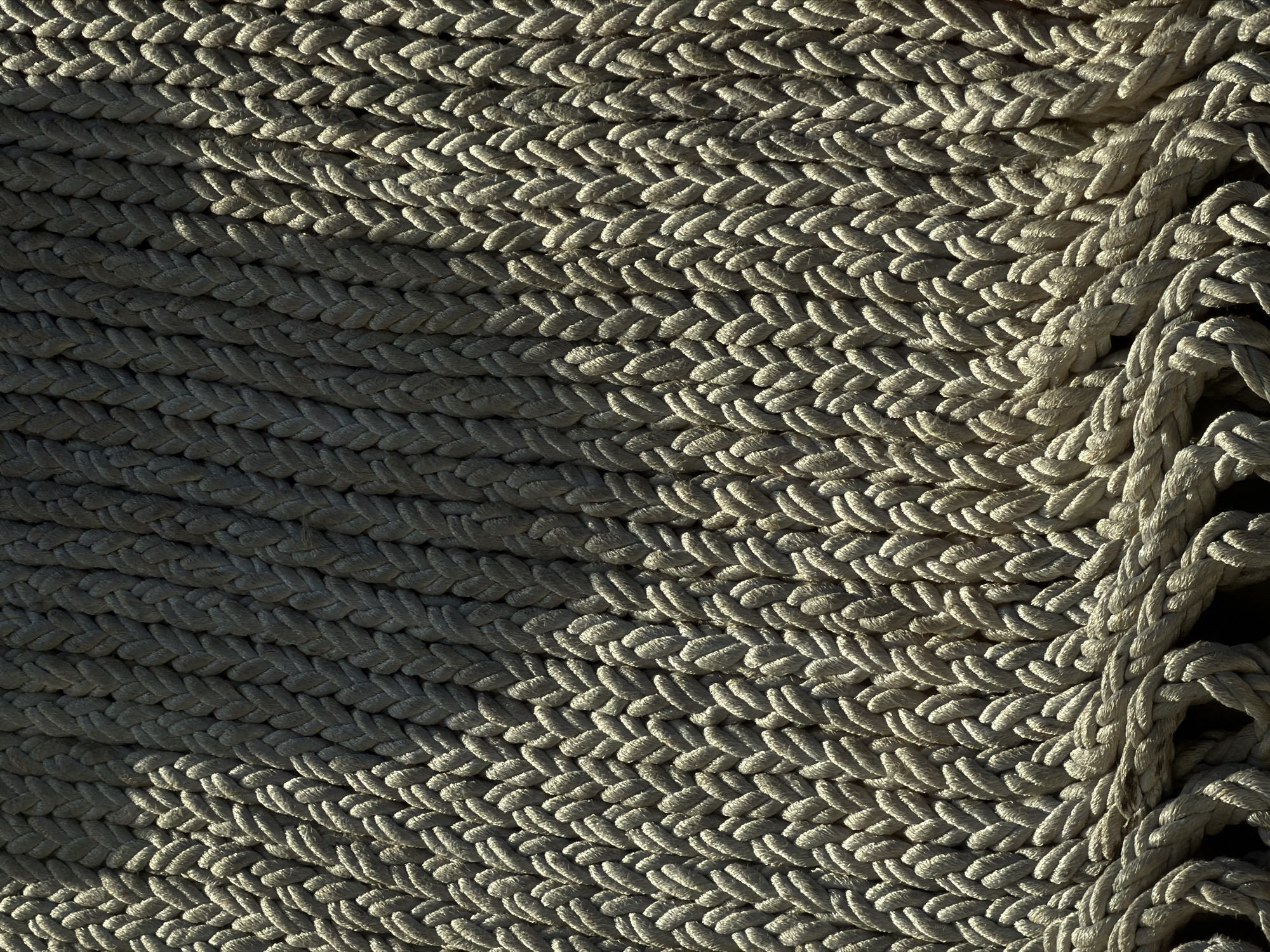 The image presents a detailed close-up of tightly coiled, light-colored rope. The rope is arranged in a consistent, repeating pattern, with the coils running parallel to each other. A shadow bisects the image diagonally, creating a contrast between a darker, shaded area on the left and a brightly lit area on the right. The texture of the rope is prominent, with individual strands visible. Some loose rope is seen on the right side of the frame. The location and city are unknown.