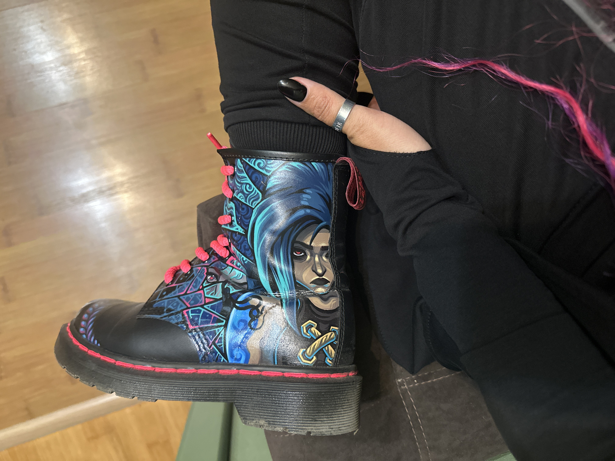 A close-up shot focuses on a black boot with custom artwork, likely in an indoor setting. The boot, possibly a Dr. Martens style, features artwork depicting a female character with blue hair and an intense expression, along with geometric and abstract designs in blue, pink, and black. The boot has pink laces and a distinctive red-stitched sole.
A person wearing black pants and a long-sleeved black shirt is holding the boot. The person has a thumb visible with a black nail polish and a silver ring. A strand of pink-colored hair is visible near the upper right edge of the frame.
The environment appears to be a room with a wooden floor visible in the background. The boot is resting on what looks like a brown fabric-covered surface.
The location in the city of Novosibirsk, Russia, cannot be confirmed from the visual information provided.