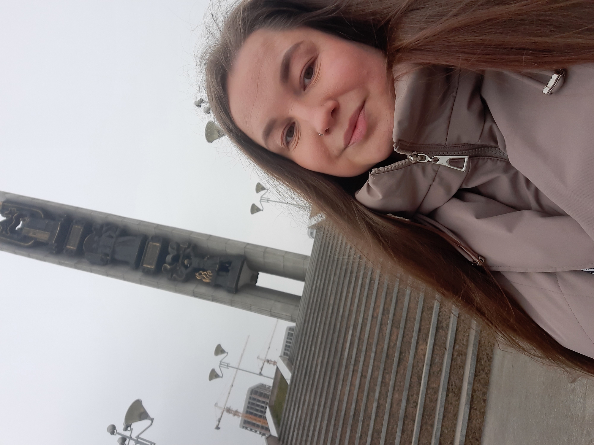 A selfie of a woman with long brown hair wearing a beige jacket. She is outside, on the steps of a monument. In the background is the "Forever with Russia" monument in Izhevsk, Russia, with additional street lighting and construction cranes in the distance. The sky is overcast.