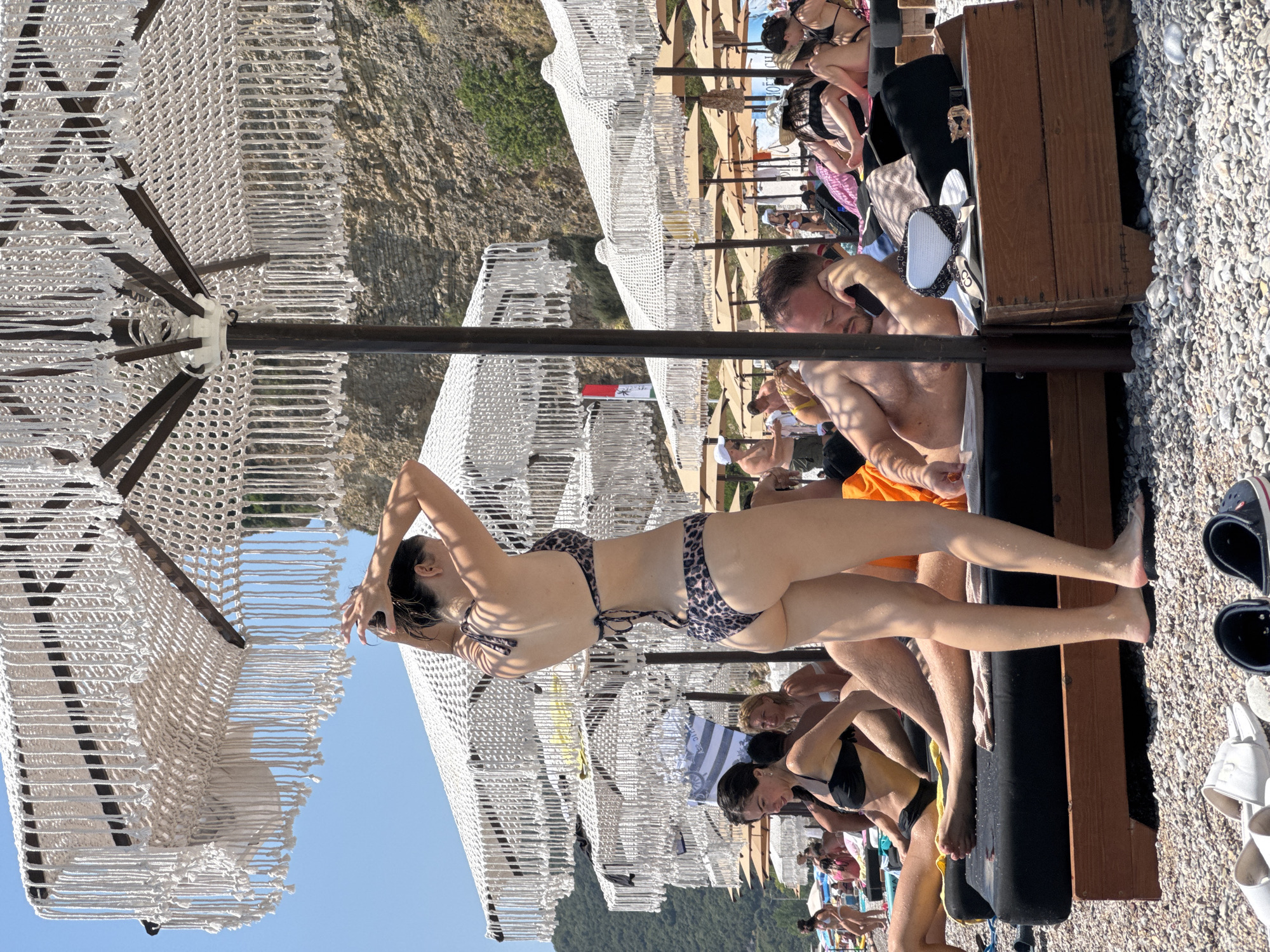 A woman in a leopard print bikini stands under a macrame-fringed beach umbrella, adjusting her hair. She is on a pebbly beach in Prijevor, Montenegro, where other beachgoers are relaxing on lounge chairs under similar umbrellas. In the foreground, a man in an orange sarong is talking on his phone, lying on a lounge chair. In front of him, a wooden box holds a hat and sunglasses. A woman in a black bikini sits on a lounge chair to his left. The beach is crowded with people, and in the background, a rocky hillside covered in greenery rises behind the umbrellas. The sky is clear and blue.