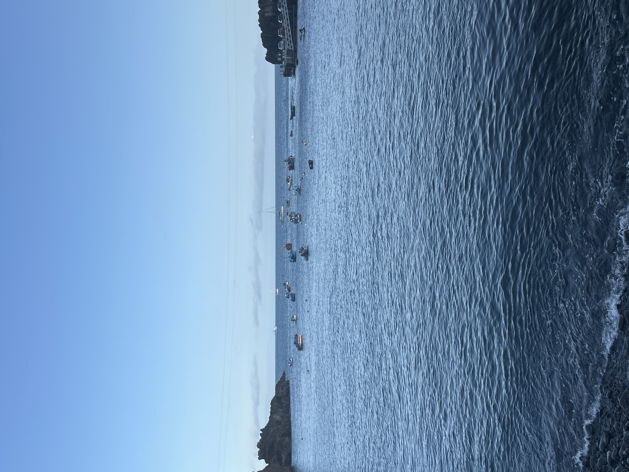 The image depicts a coastal scene, with a serene view of the sea and sky, likely in the city of Câmara de Lobos, Portugal. The sky is clear with shades of blue, suggesting a bright day. The calm water is populated with several boats, some of which are sailing vessels, moored or anchored in the bay. The shoreline is partially visible on either side of the image, composed of rocky cliffs and a harbor. There are electrical wires visible near the horizon.