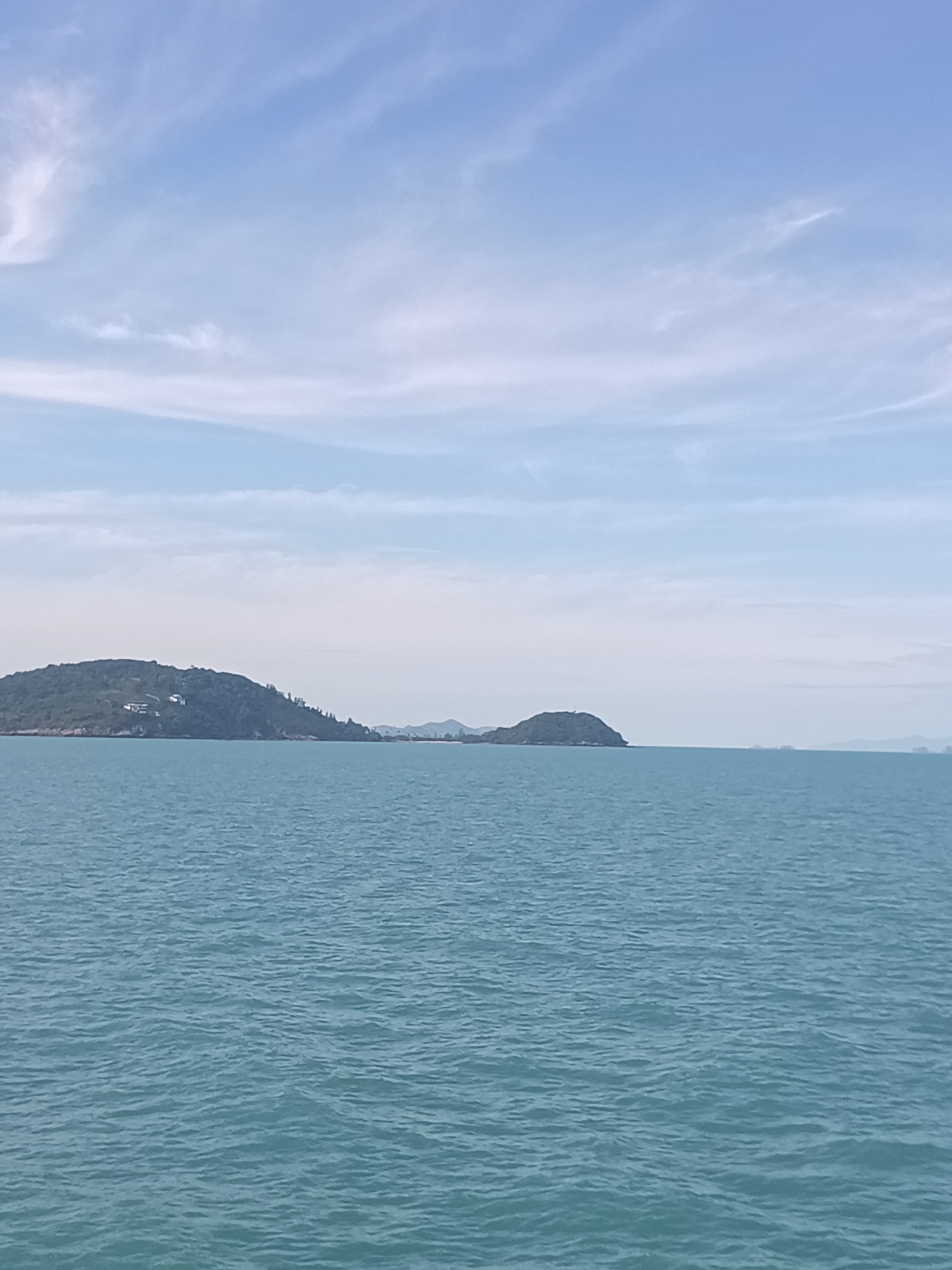 The image depicts a tranquil seascape near Baan Thong Po, Thailand. The foreground is dominated by turquoise water with gentle ripples. In the midground, two islands are visible. The larger island on the left is lush green with some white structures. A smaller island is to the right. The background is a pale blue sky with wispy clouds.