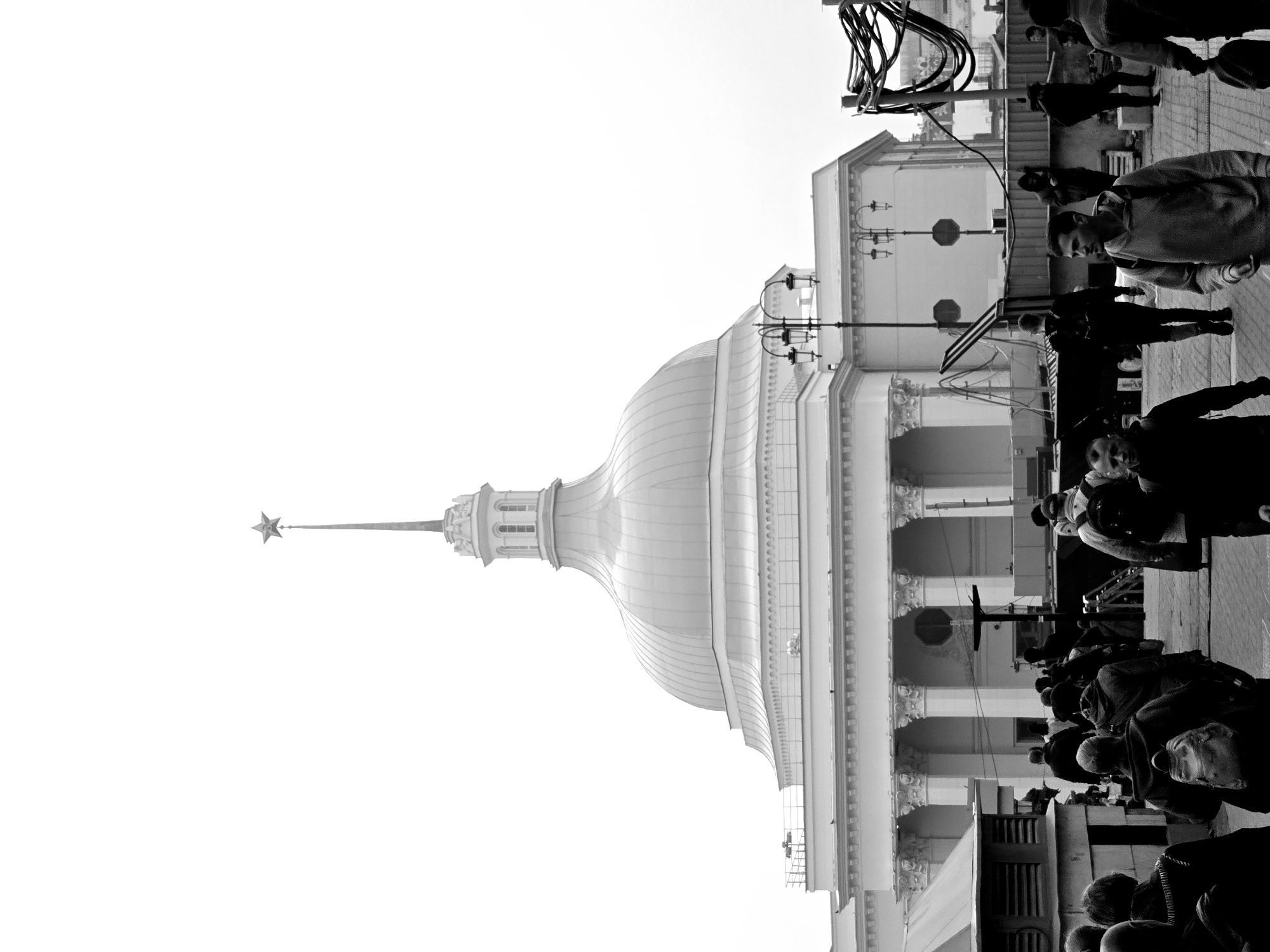 The image is a black and white photograph of a building in Moscow, Russia. The building has a large dome topped with a star. A crowd of people is present in front of the building.