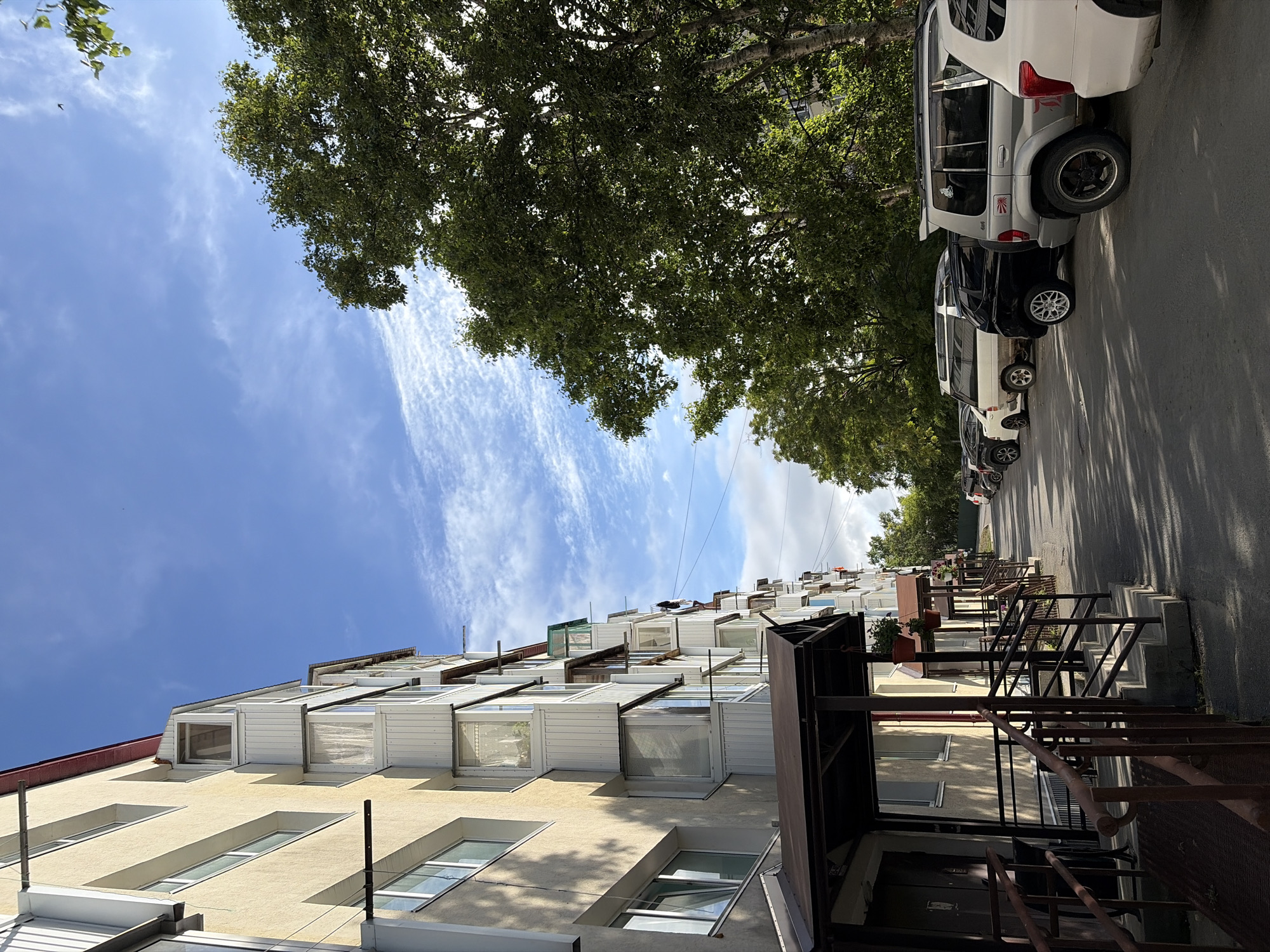 This image captures a street scene in the city of Korsakov, Russia, on a clear day with scattered white clouds in a bright blue sky. In the foreground, the side of a light yellow apartment building is visible, characterized by its numerous windows and protruding balconies, some of which are enclosed with white paneling and curtains. A covered walkway with a dark brown roof extends along the building's entrance, with concrete steps and metal railings leading up to the doorways.
To the right of the apartment building, a row of parked cars lines the asphalt street. Among them are several SUVs, including a silver SUV with a red decal on its rear panel and a white SUV at the far right. The cars are positioned parallel to the building. A dense canopy of green trees with a textured bark, likely birch trees, forms a backdrop on the right side of the image, partially obscuring other buildings and structures in the distance. The lighting suggests it is daytime, with shadows cast on the street and the side of the apartment building.