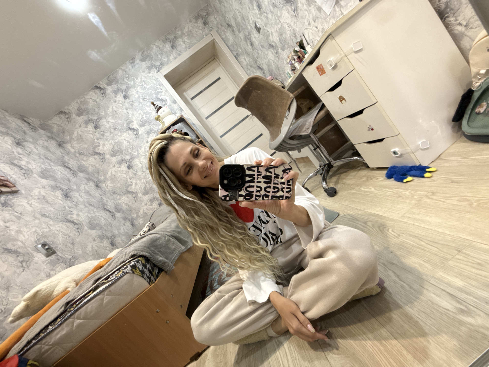 A woman with blonde dreadlocks is taking a selfie in what appears to be a room in a home in Rusanovo, Russia. She is sitting cross-legged on a light-colored wooden floor wearing a white shirt with a large red heart graphic, and light beige sweatpants. She is holding a phone with a patterned case in front of her face, presumably taking the photograph.
Behind her is a bed with various pillows and blankets. The room has grey marble-patterned wallpaper. Further in the background, there is a desk with white drawers, an office chair, and a white door with horizontal panels. A blue toy is visible on the floor near the desk. The lighting in the room is bright.