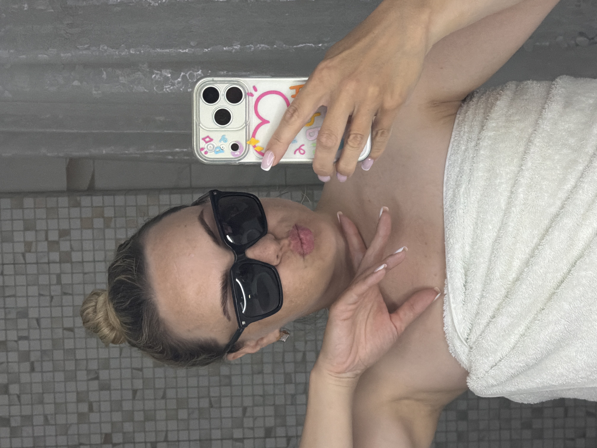 A woman wearing sunglasses and a white towel is taking a selfie in a bathroom. Her hair is pulled back in a bun. She is holding a phone with a decorated phone case. The bathroom has gray tiled walls and floor. There is no information to suggest that this image was taken in the city of Votkinsk, Russia.