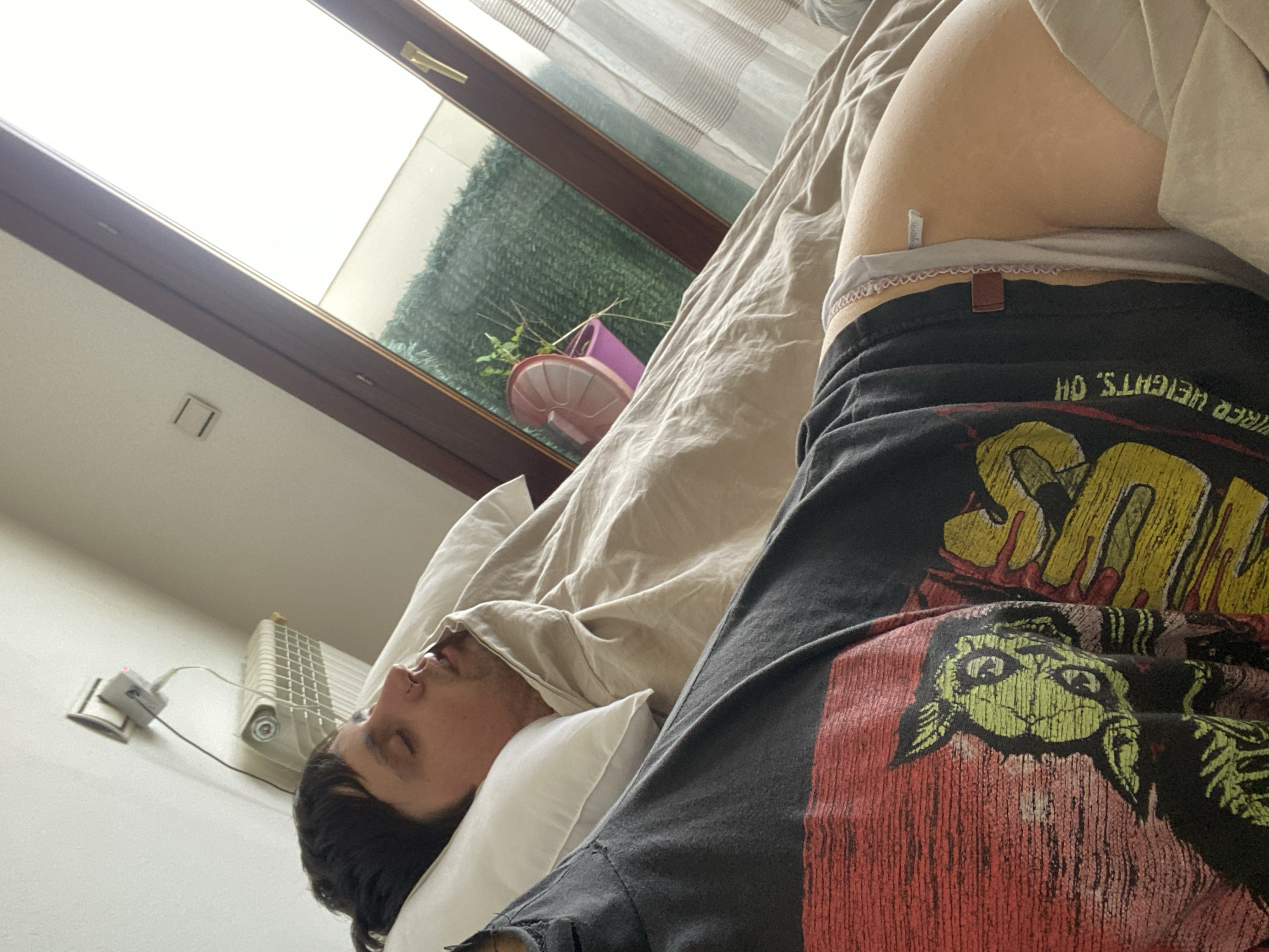 The photograph shows a man sleeping in a bed. He is lying on his back with his head on a white pillow and covered partially by a beige blanket. A window with a wooden frame is visible in the background, through which greenery and a pink pot can be seen. To the left of the man, there is a white radiator and an electrical outlet. A person wearing dark-colored pants and underwear is sitting nearby. The image was likely taken indoors in a residential setting. While the exact location within Gijón/Xixón is not discernible from the image, the architectural style and presence of indoor plants suggest a domestic environment that could be found in many parts of the city.