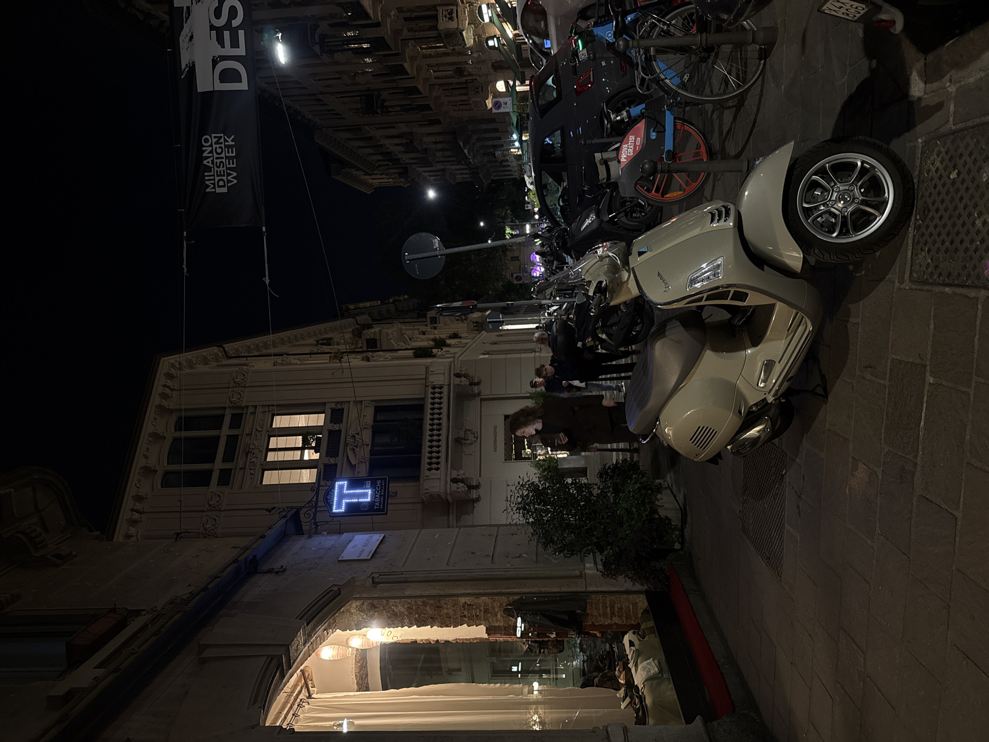 The image shows a street scene at night in Milan, Italy. A beige scooter is parked on the cobblestone sidewalk. Parked bicycles are visible along the street in the background. A banner with "MILANO DESIGN WEEK" is hanging on a building. People are standing and walking on the sidewalk. A blue neon sign with a "T" indicates a tobacco shop (TABACCHI). A restaurant or store with lit displays is seen through an arched entrance. The buildings are ornately decorated with architectural details.
