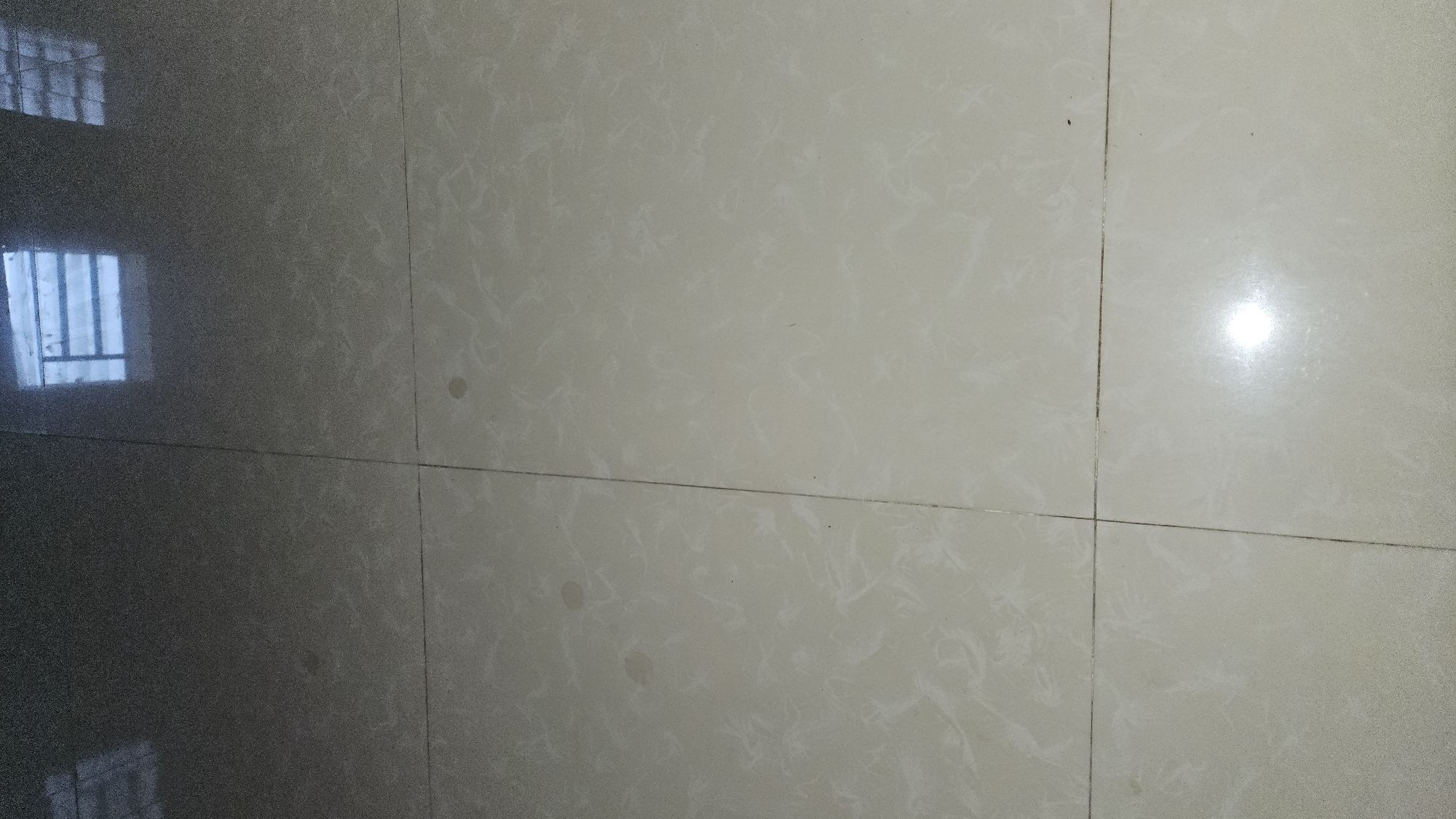 The image shows a section of tiled flooring. The tiles appear to be light-colored with a subtle mottled pattern. Grout lines are visible between the tiles. On the left side of the image, there is a dark reflection of what appears to be a window with bars. The reflection indicates the surrounding environment may be in Cam Đức, Vietnam. The flooring is clean and uncluttered. There are no people or other objects present.