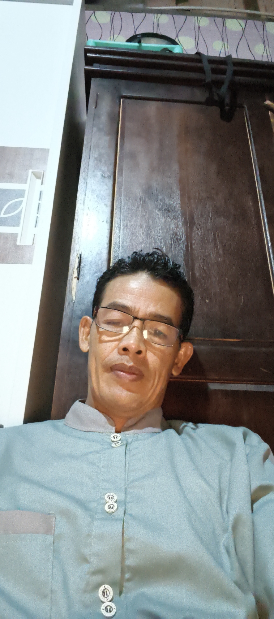 A selfie taken indoors, likely in Muaro Jambi, Indonesia. The subject, a man wearing glasses and a light blue collared shirt with decorative buttons, is positioned in the lower half of the frame, looking directly at the camera. His face is illuminated from the front, casting subtle shadows. Behind him is a large, dark wooden cabinet with a patterned wallpaper above it, featuring light-colored circles and dark lines. To the left of the cabinet is a section of a white wall with a decorative element. The lighting suggests an indoor setting, possibly a home or a personal space, with the light source originating from the front and slightly to the left of the subject.