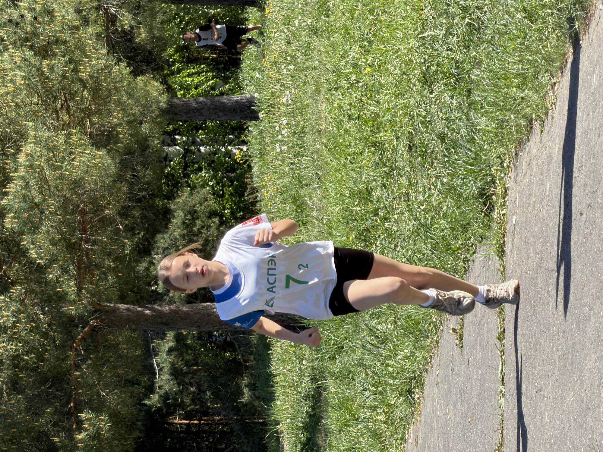 A young woman is running on a paved path next to a grassy area with scattered flowers and a tree line. She is wearing a white t-shirt with the word "АСПЭК" and the number "72" on it, along with black shorts and running shoes. In the background, another person can be seen running in the same direction. The location appears to be Забегалово, Russia, based on the shirt's details. The scene is captured in daylight.