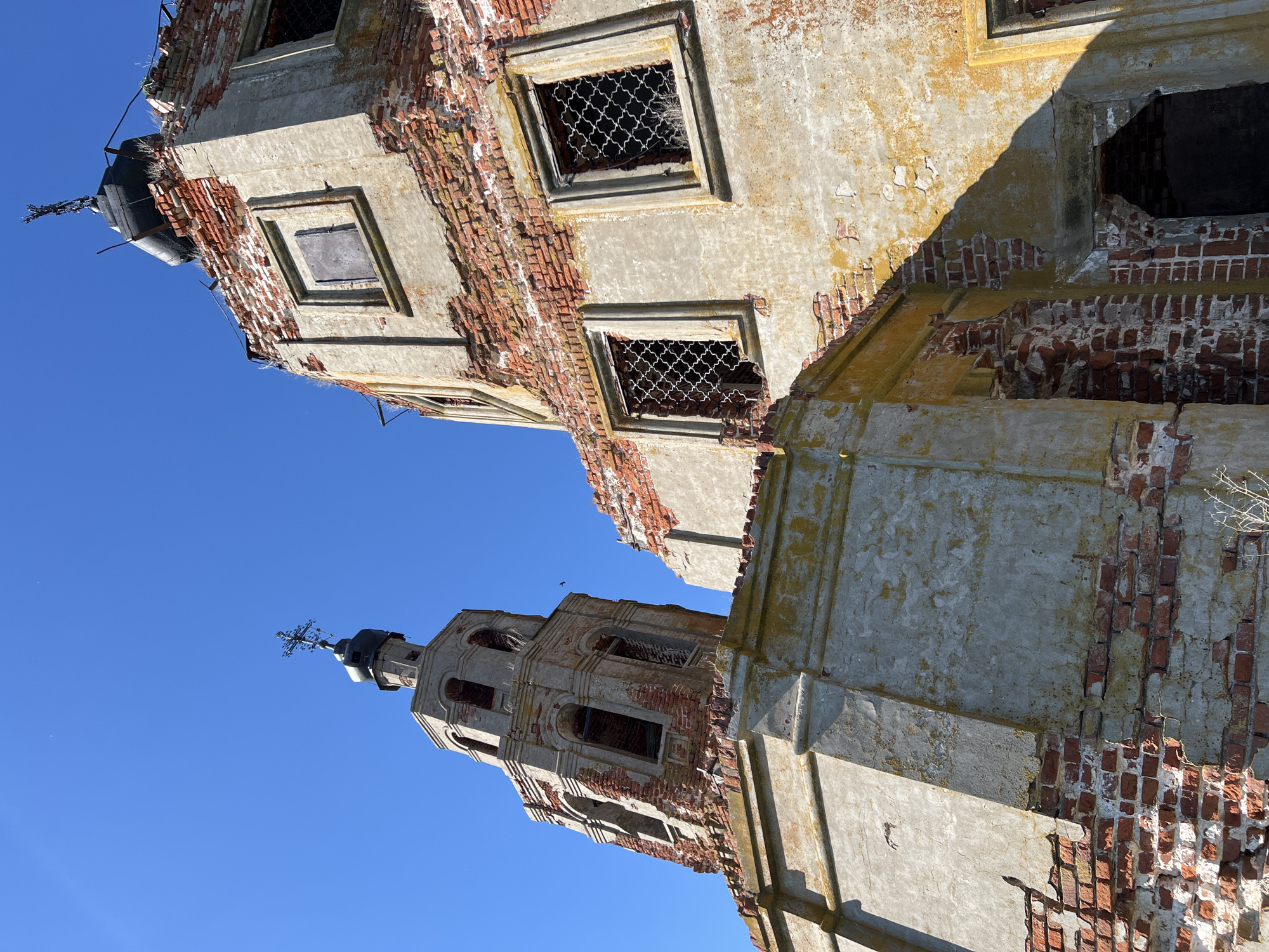 The image depicts the decaying remains of a brick and concrete structure, possibly a church, in Шуран, Russia, under a clear blue sky. The structure is heavily weathered, with crumbling brickwork and dilapidated walls. Window openings are visible, some with remnants of iron grates. A tower topped with a cross is present. The image is shot from a low angle, emphasizing the height and decay of the structure.